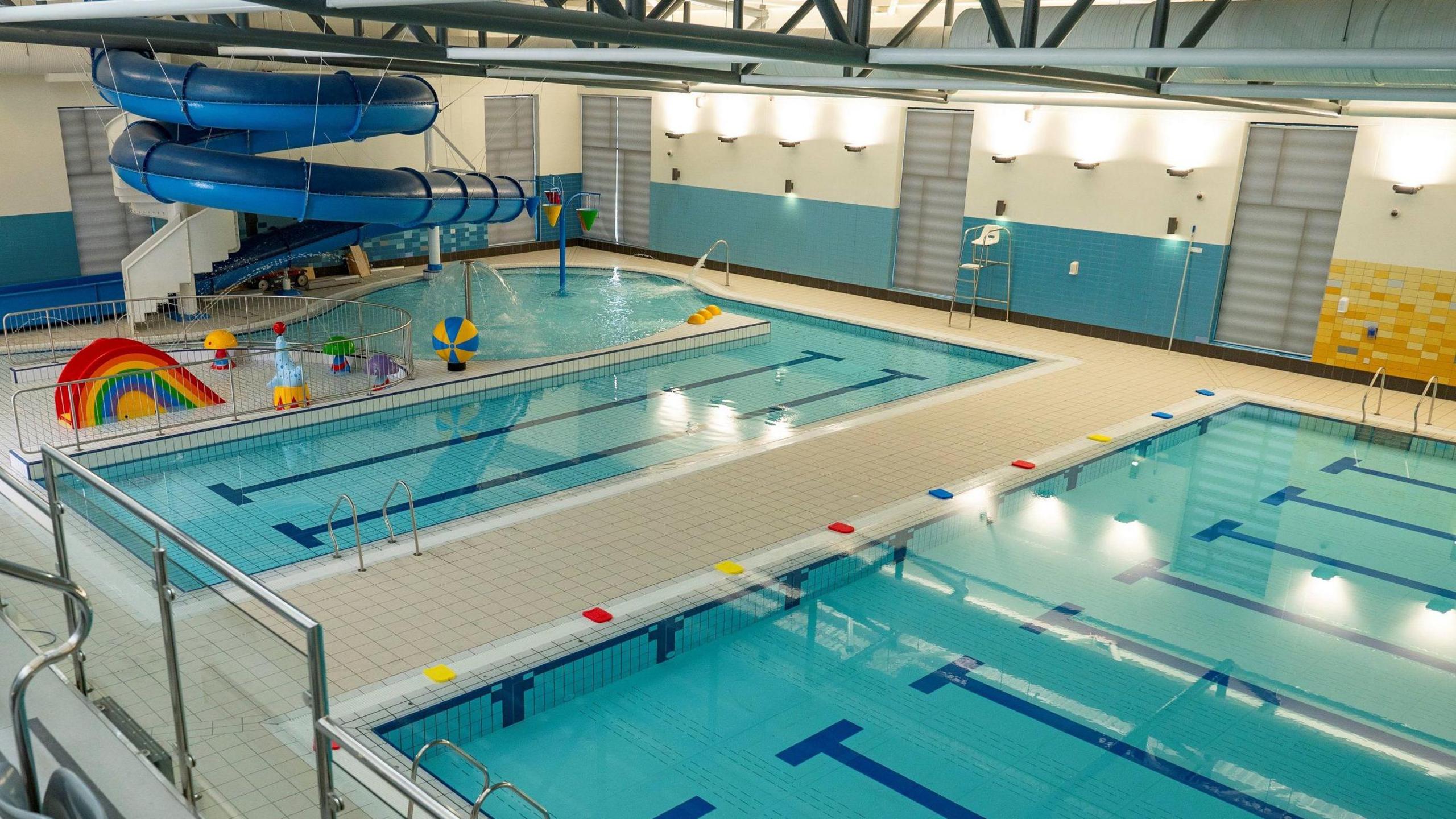 An aerial view of Newry Leisure Centre's pool hall taken from its viewing gallery.  There is a 25m main pool, a smaller learner pool and a children's play area.  A large blue flume curls down from just below the ceiling. 