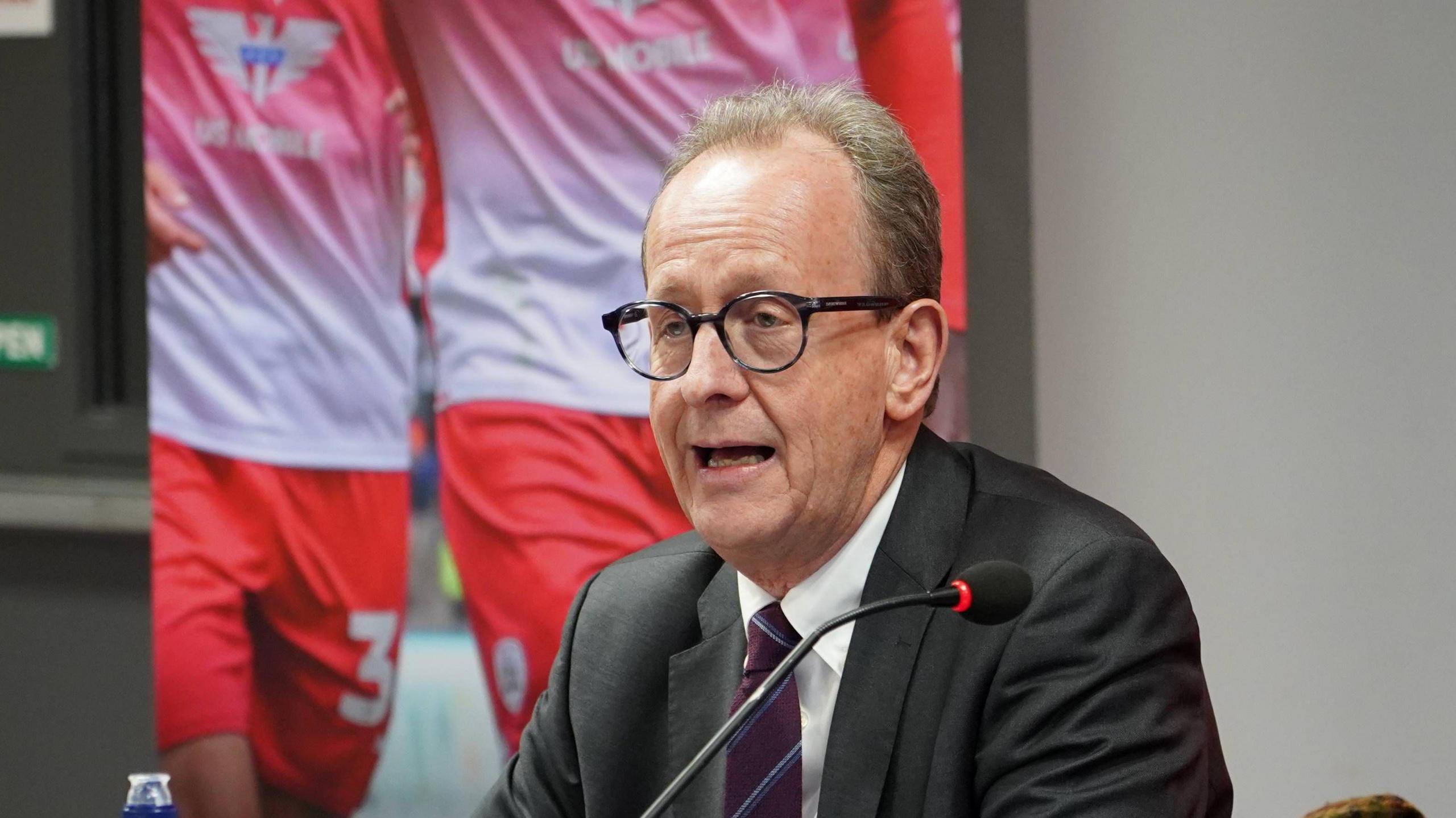 Barnsley Council leader Sir Stephen Houghton is in a grey suit with a striped burgundy and blue tie and wearing glasses. He is sat in front of a microphone. Behind him is a picture showing two people in red football kits.