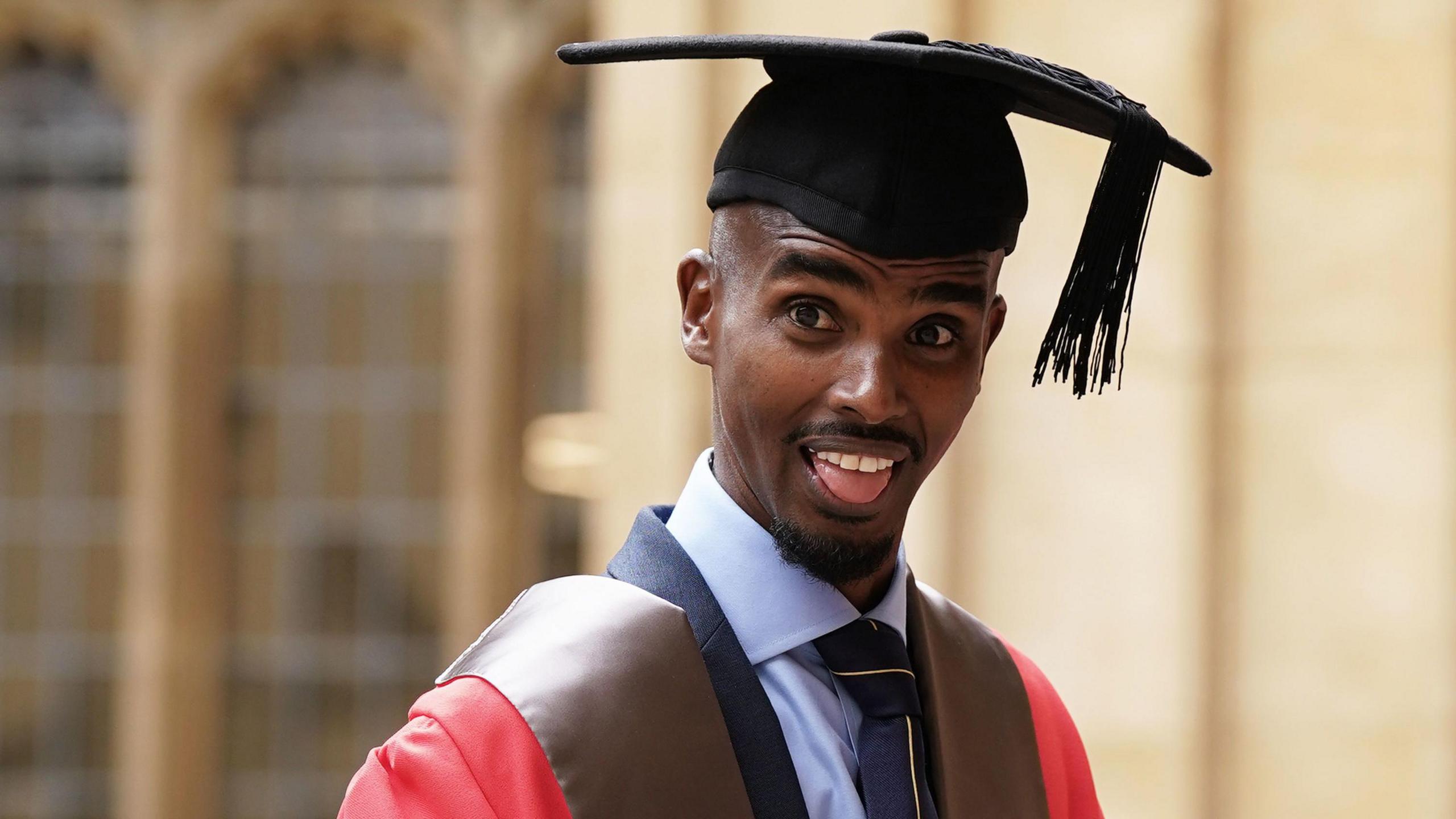 Sir Mo Farah has his tongue out, and is wearing a red gown and black square cap.