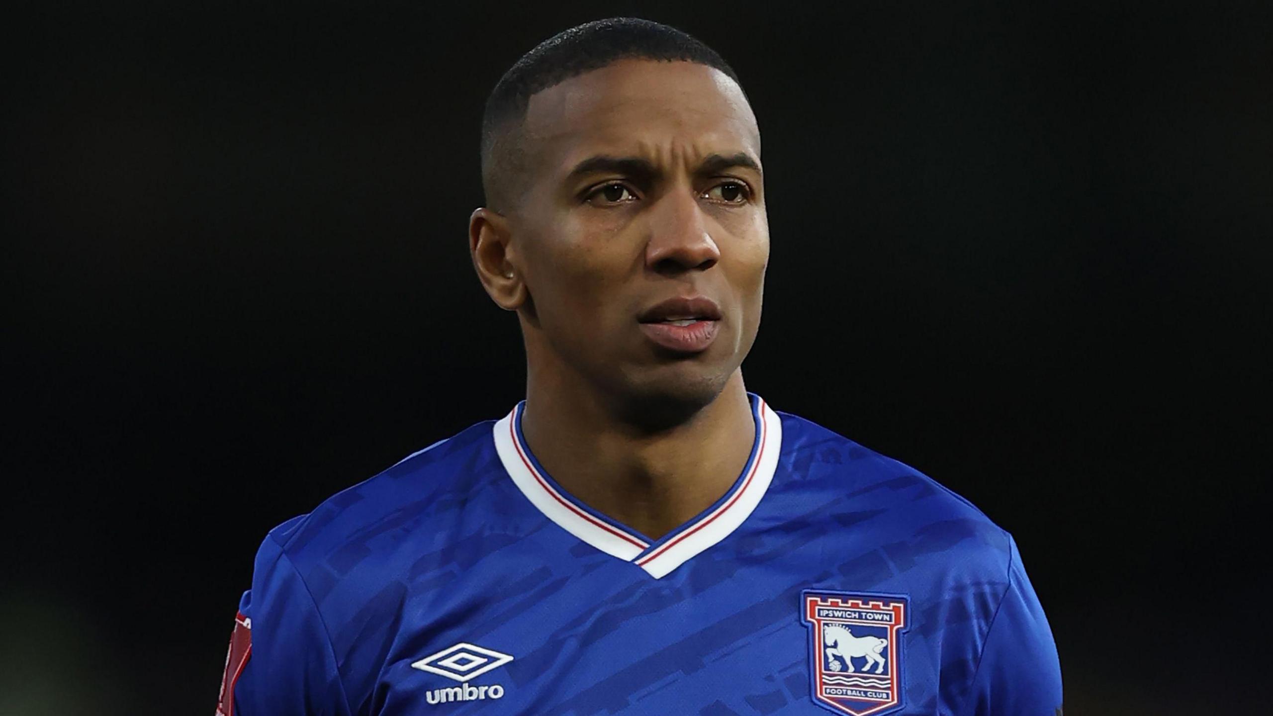 Ashley Young, wearing Ipswich Town's blue home shirt with a white collar and red and blue trim. The shirt has an Umbro logo on one side and Ipswich crest on the other.