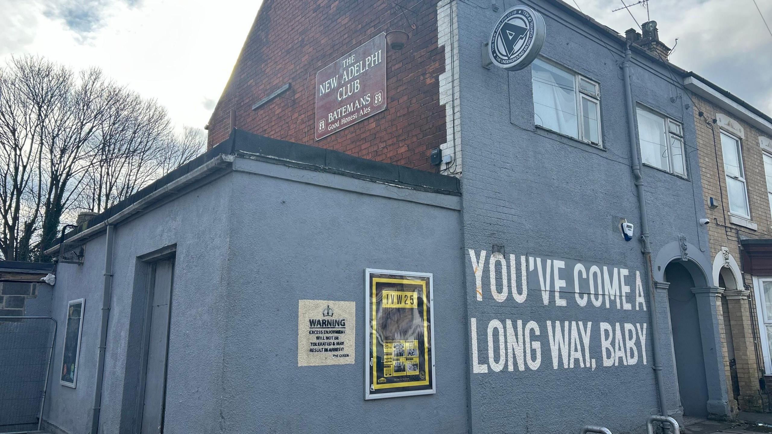 The exterior of the New Adelphi Club. The photo shoes a grey-painted building with a red-brick upper section. A large mural on the wall reads, "YOU'VE COME A LONG WAY, BABY." There's a warning plaque and a framed poster near the entrance.