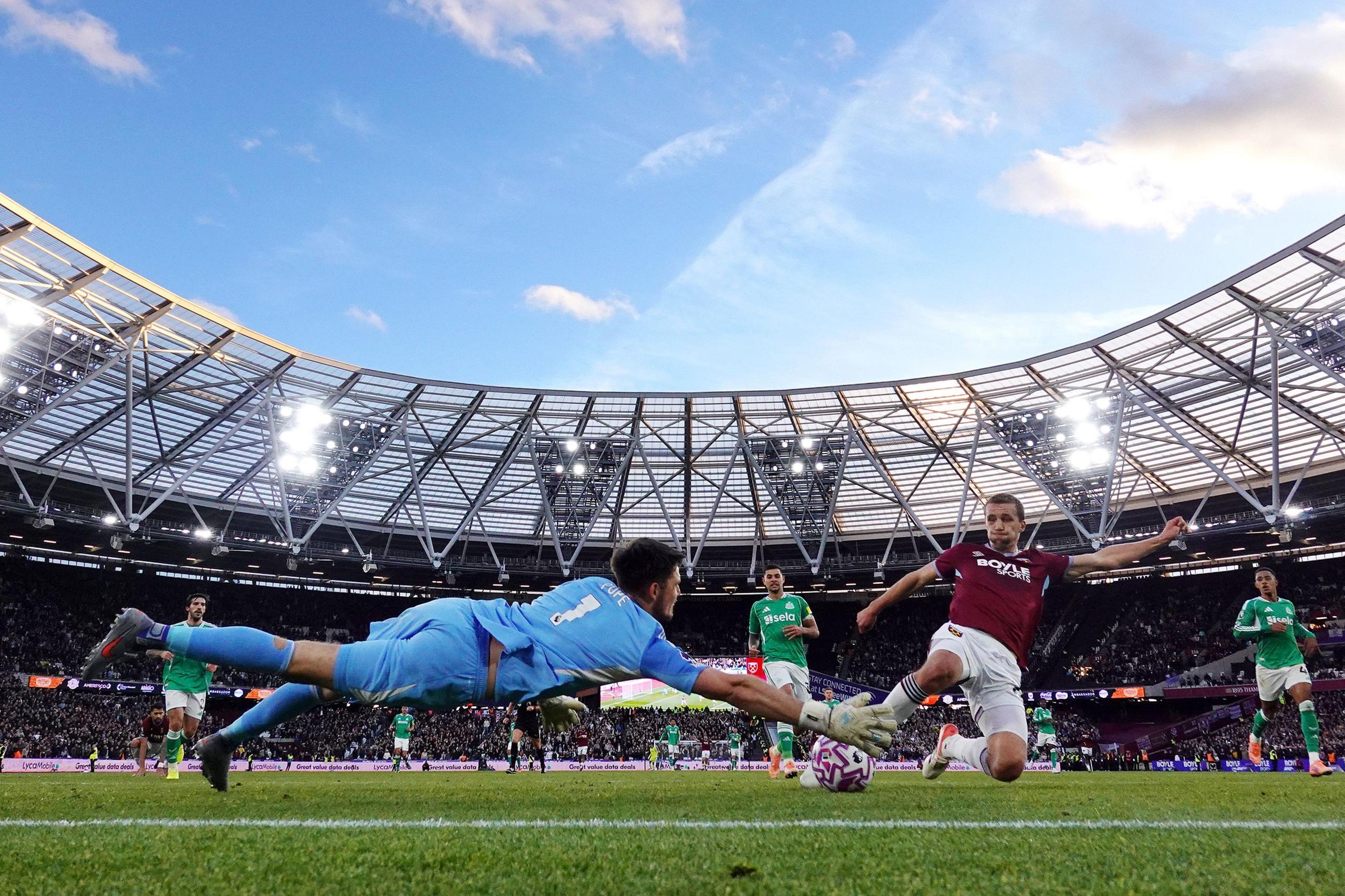 A goalkeeper in a light blue kit dives fully extended toward the ball near the goal line as an opposing player in a maroon jersey stretches a leg to shoot. Other players and a packed stadium under a bright sky form the backdrop.