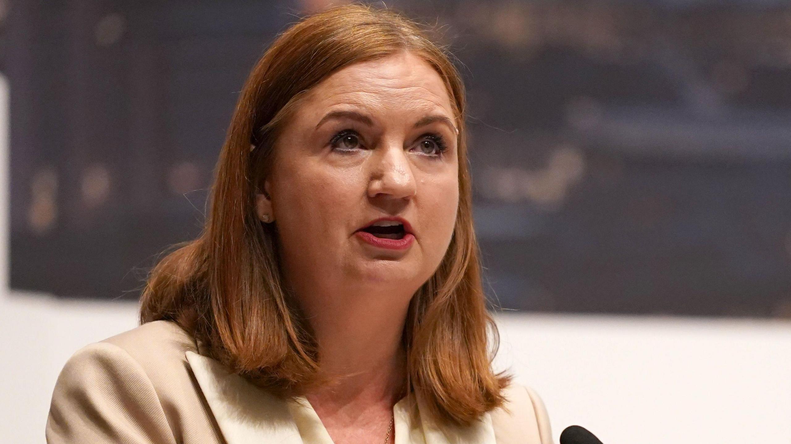 Jayne Brady has short red/brown hair and is speaking into a microphone. She is wearing a beige suit.