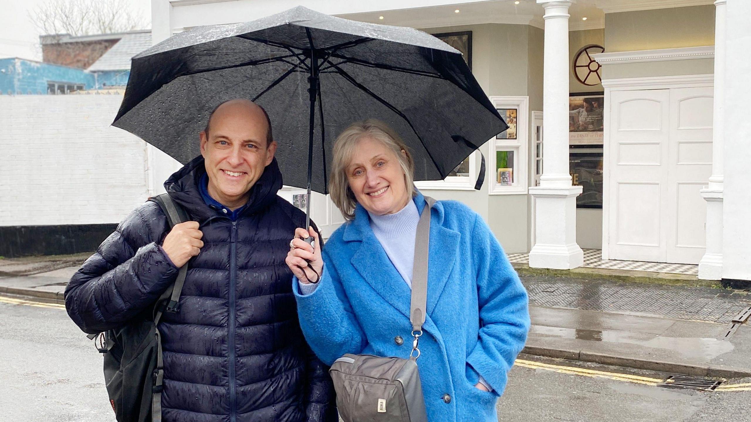 Christopher Baines and Helen Sheppard smiling for the camera outside, while it's raining. She is holding a black umbrella. They are both wearing thick coats. The street behind them is wet.