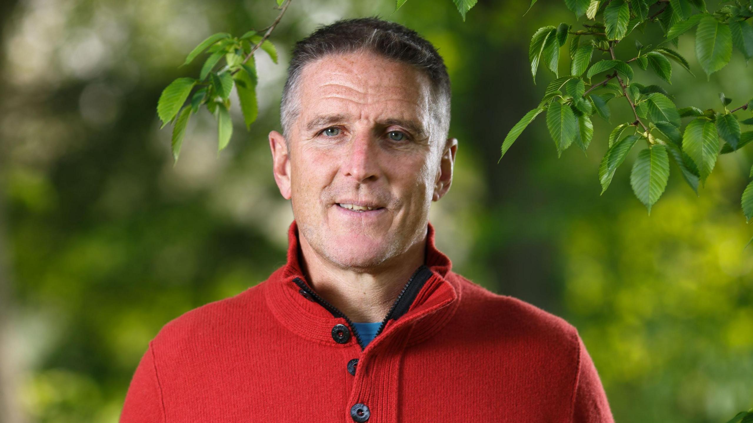 Iolo is wearing a bright red knitted jumper with black buttons. He is looking at the camera and has short grey hair.