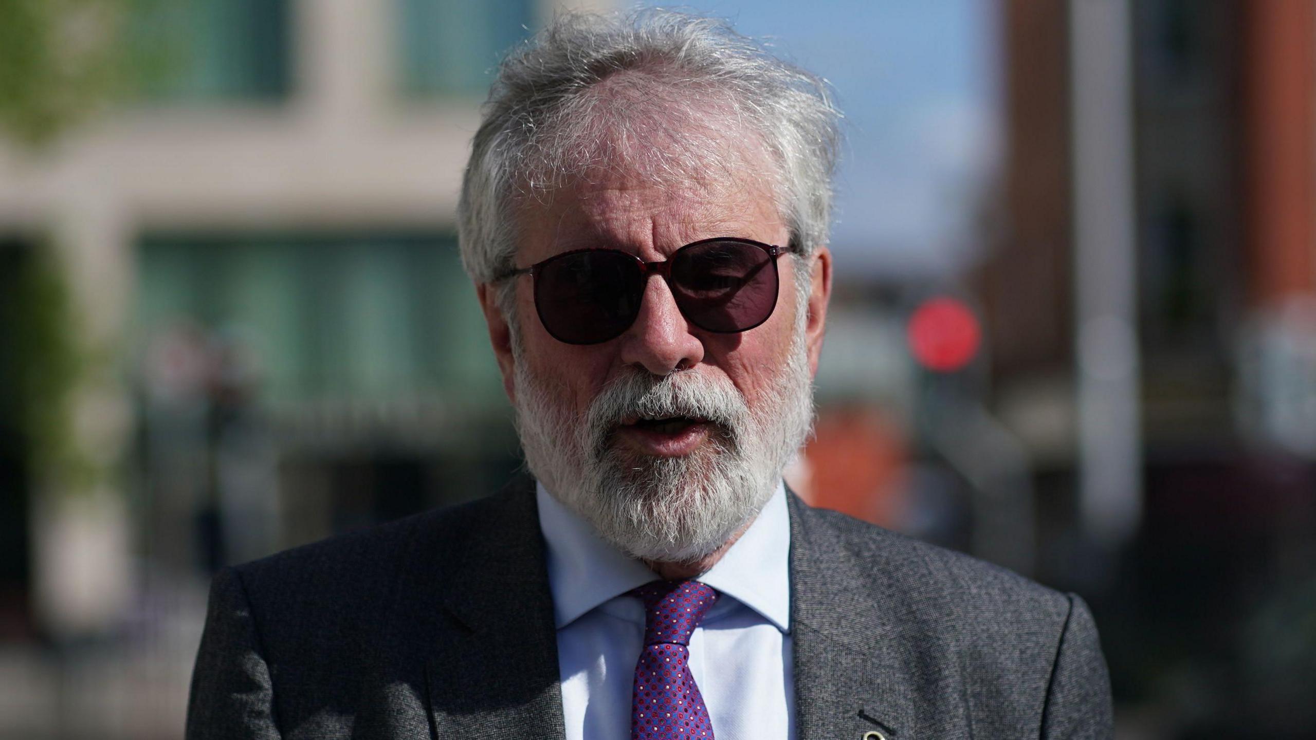 Gerry Adams, wearing a grey suit jacket, a light coloured collared shirt and purple tie. He has grey hair and a grey beard and wears dark sunglasses. He is walking on the street outside of the High Court, but the background behind him is blurred.