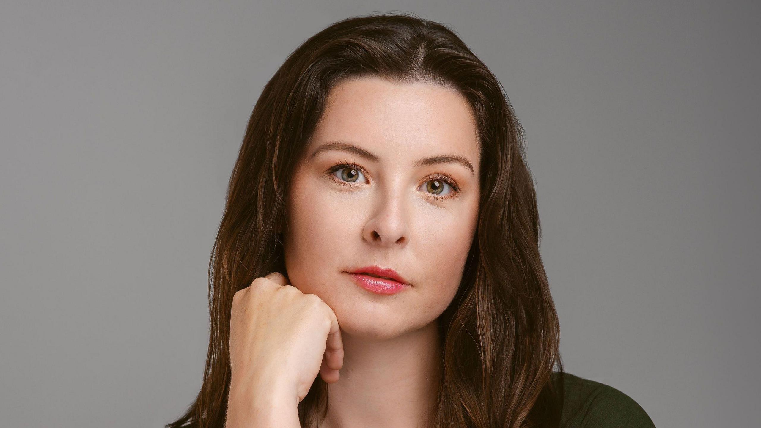 Alexa Morden, a woman with long brown hair, wearing glossy lipstick, looking into the camera with one hand resting on her chin