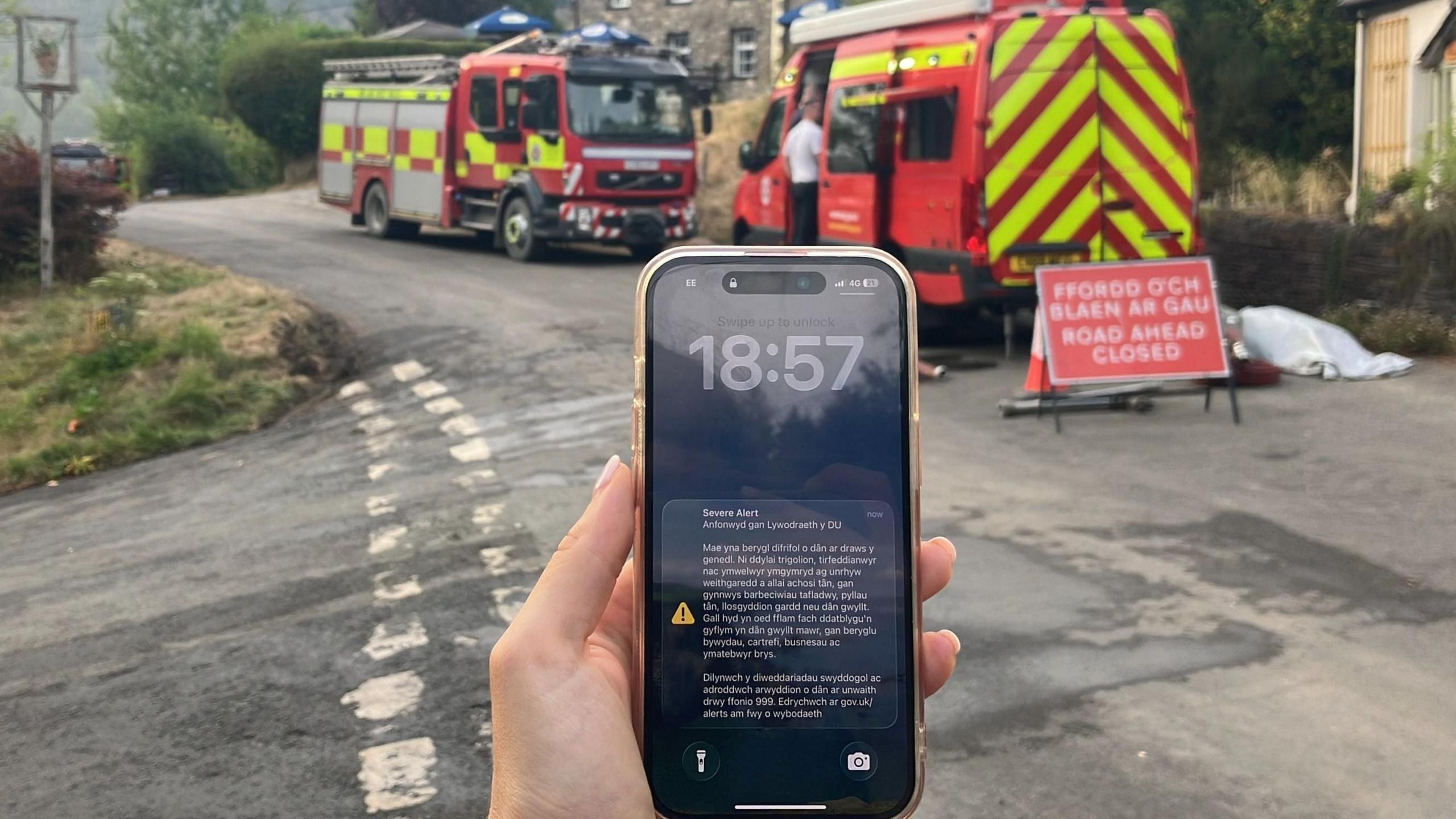 A phone screen with an emergency alert on is seen on a phone, with fire engines behind