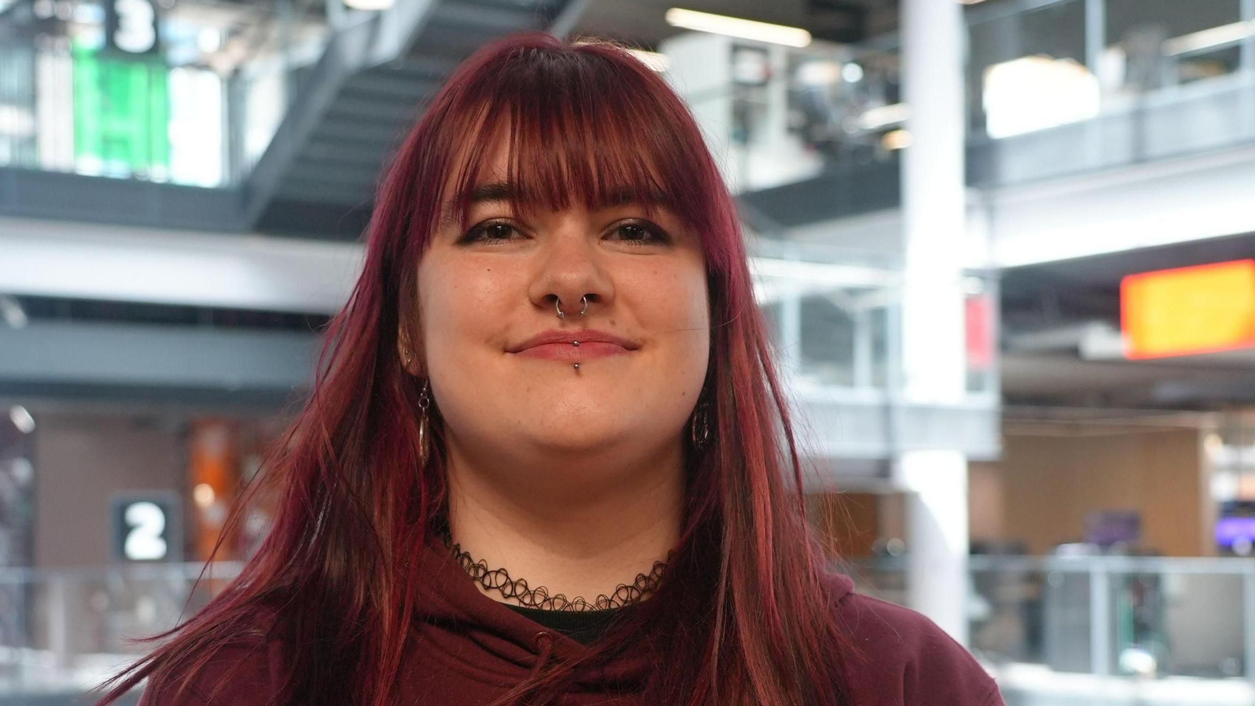 Kady is smiling at the camera. She's wearing a burgundy hoodie and has dyed red hair which is down to her shoulders. She has a nose ring and lip piercings.