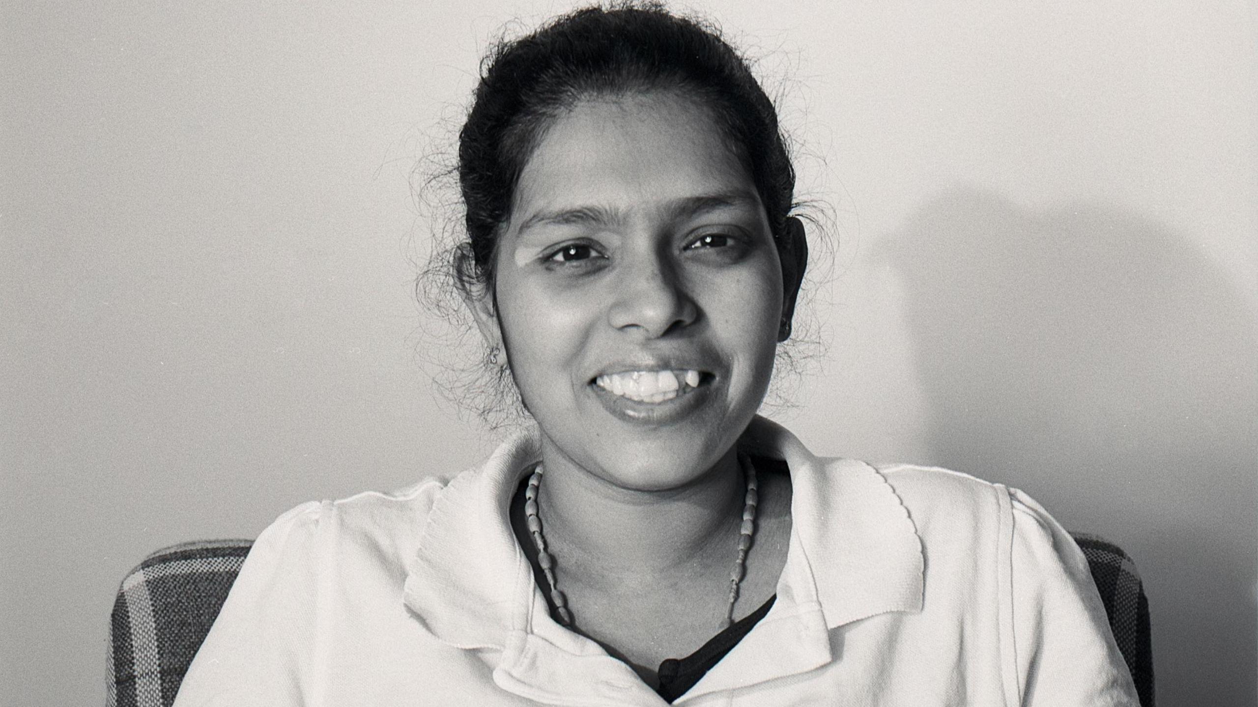 Black and white portrait of Cynthia Fernandez. She has dark hair which is tied up and is wearing a beaded necklace and shirt with a collar. She is sat in a chair against a solid background, looking directly at the camera and smiling. 