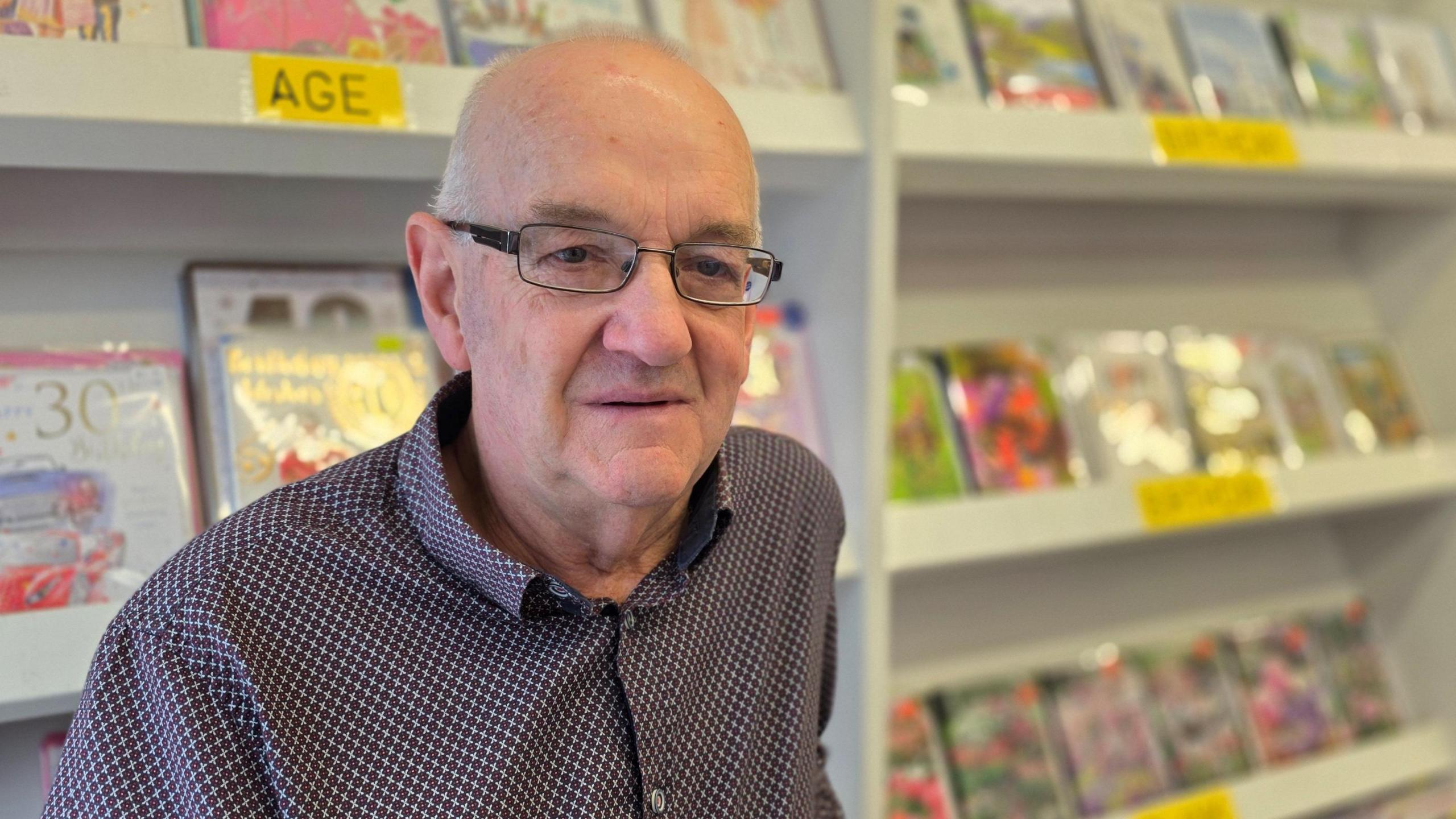 Ray Harvey is wearing a patterned shirt, with small red, black and white checks on it and standing in front of a display of greetings cards.