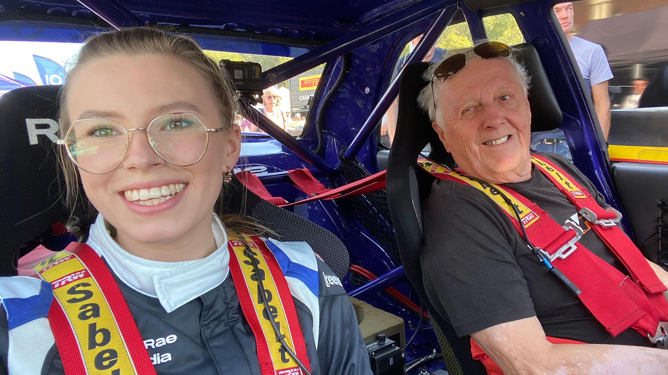 Hollie and grandfather Jimmy in a rally car.  She's wearing large, round, gold-rimmed glasses and a racing suit with a white collar and black and blue accents. A red and yellow racing harness is securely fastened across her chest. A small action camera, possibly a GoPro, is mounted on the roll cage above her. He is wearing sunglasses perched on his head. He's also wearing a racing harness, and a black T-shirt. 
