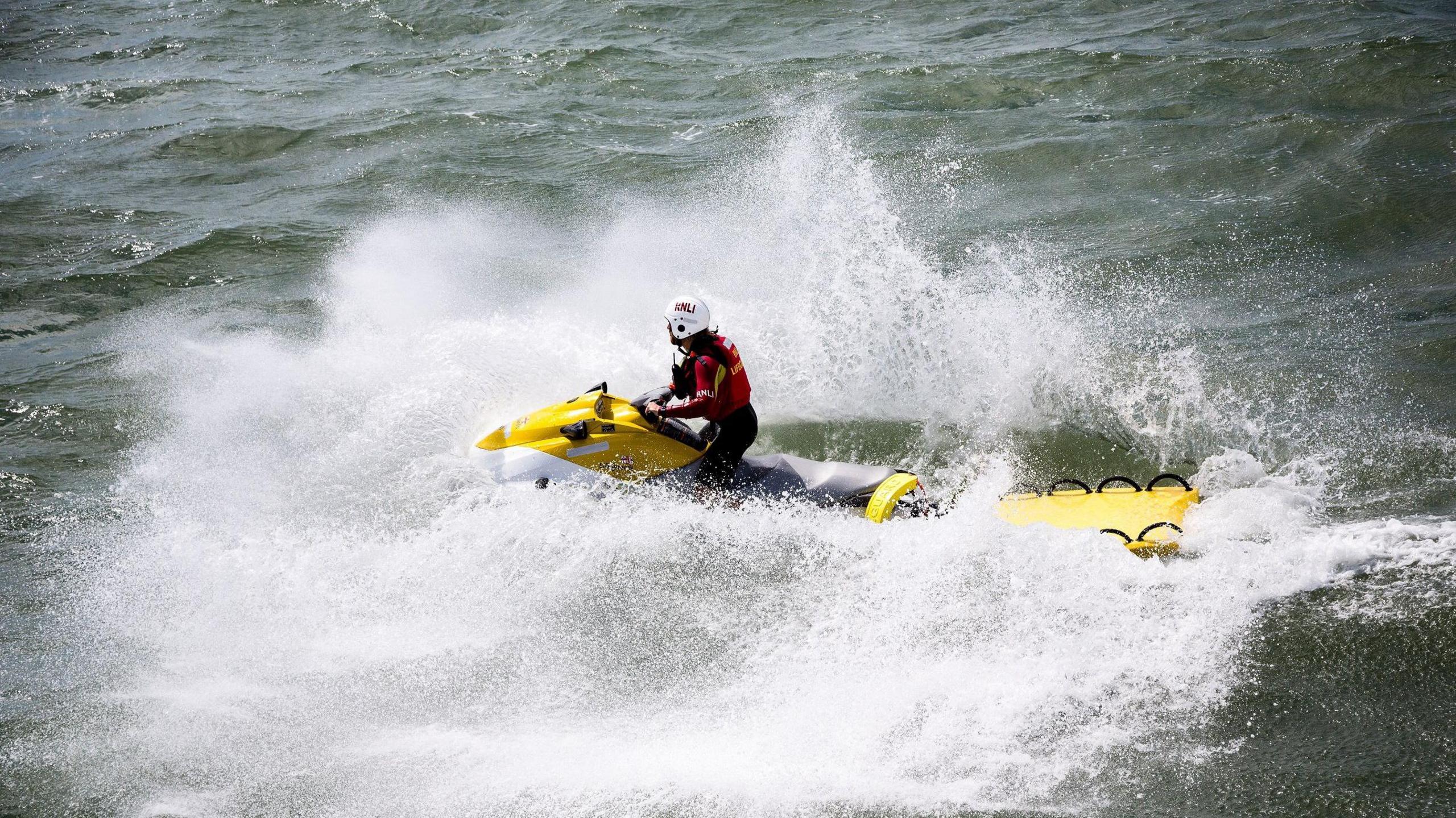 A person in RNLI uniform on a jet ski towing an empty stretcher. The sea is quite choppy and the jet ski has created a lot of wash.
