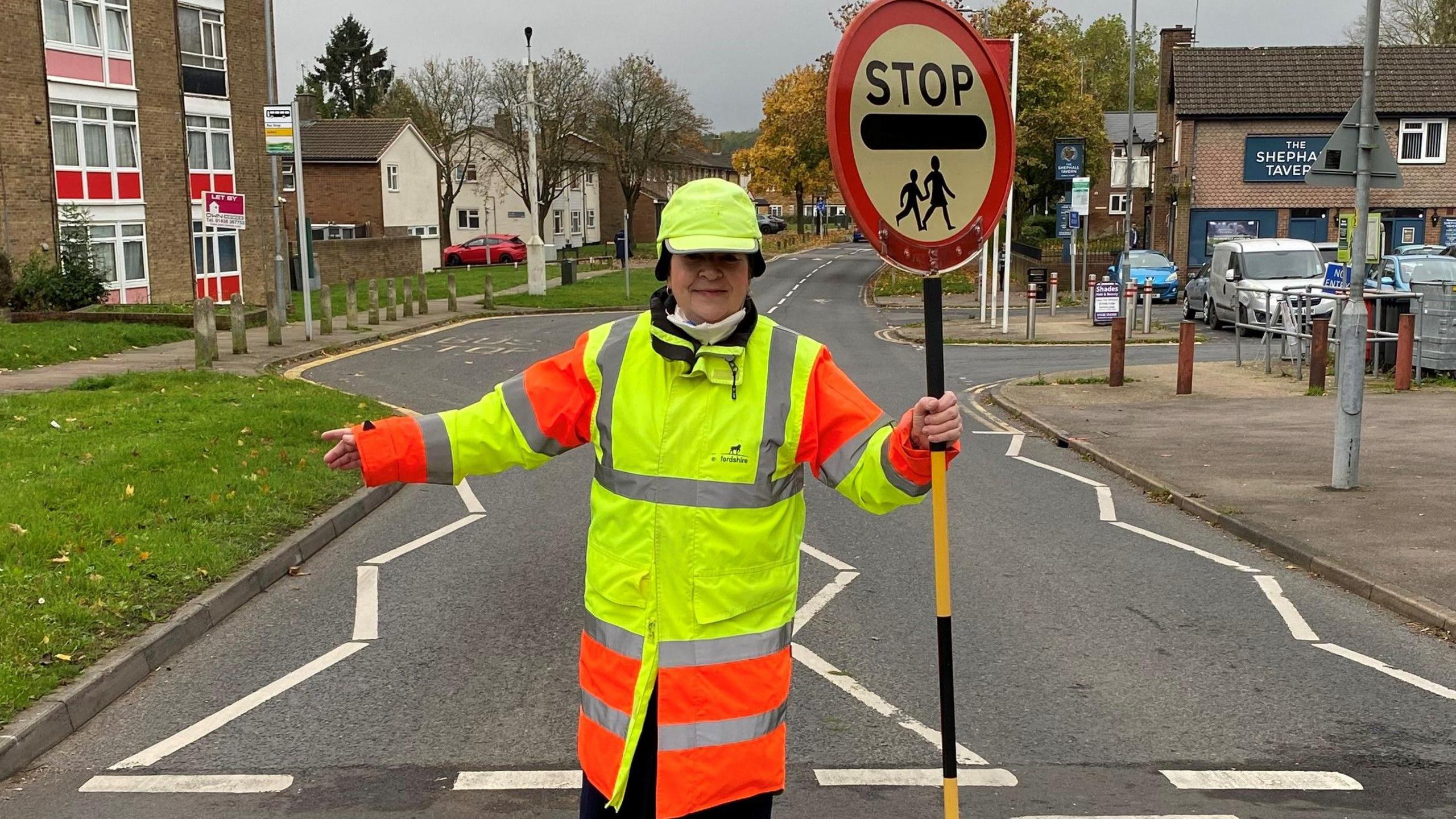 Julie Bennett, dressed as a lollipop lady, with a high vis hat, jacket and holding a lollipop patrol stick with STOP on it. She is standing in the middle of a road crossing, with one of her arms outstretched and the other holding her stick. 