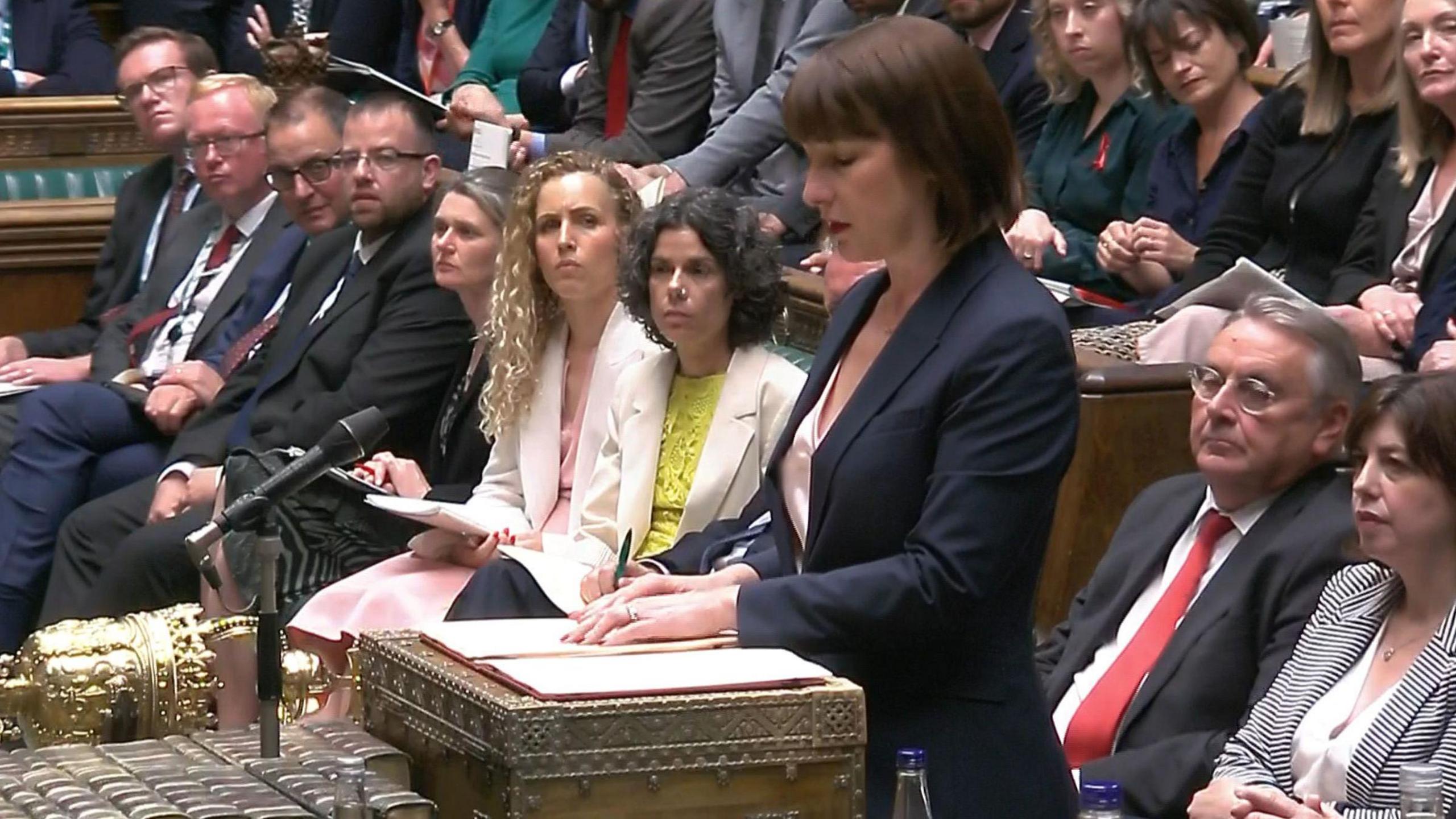 Rachel Reeves standing in Parliament surrounded by MPs who are watching her from the benches