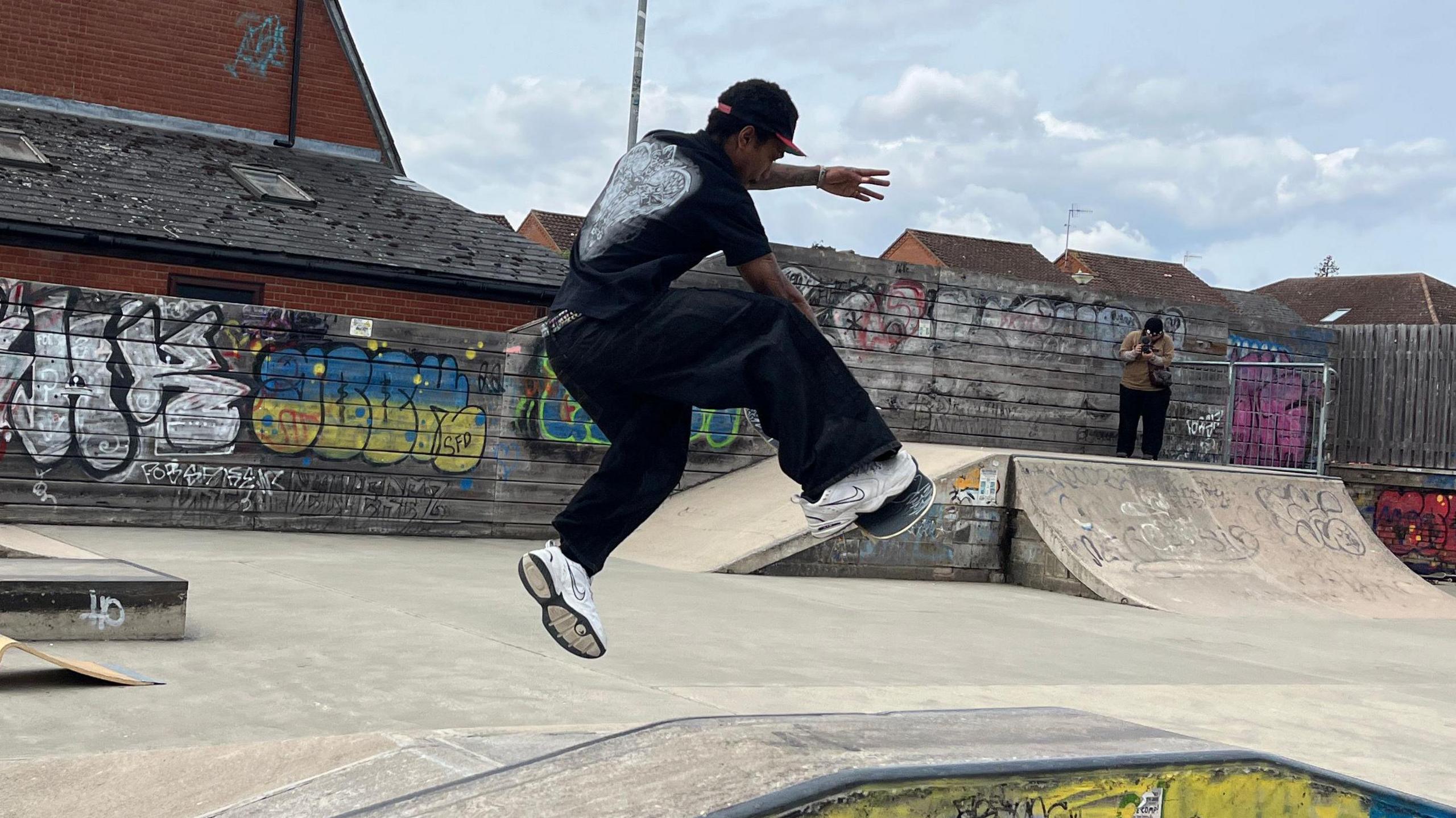 A man dressed in all black with white trainers who is skateboarding over a ramp at a skate park.
