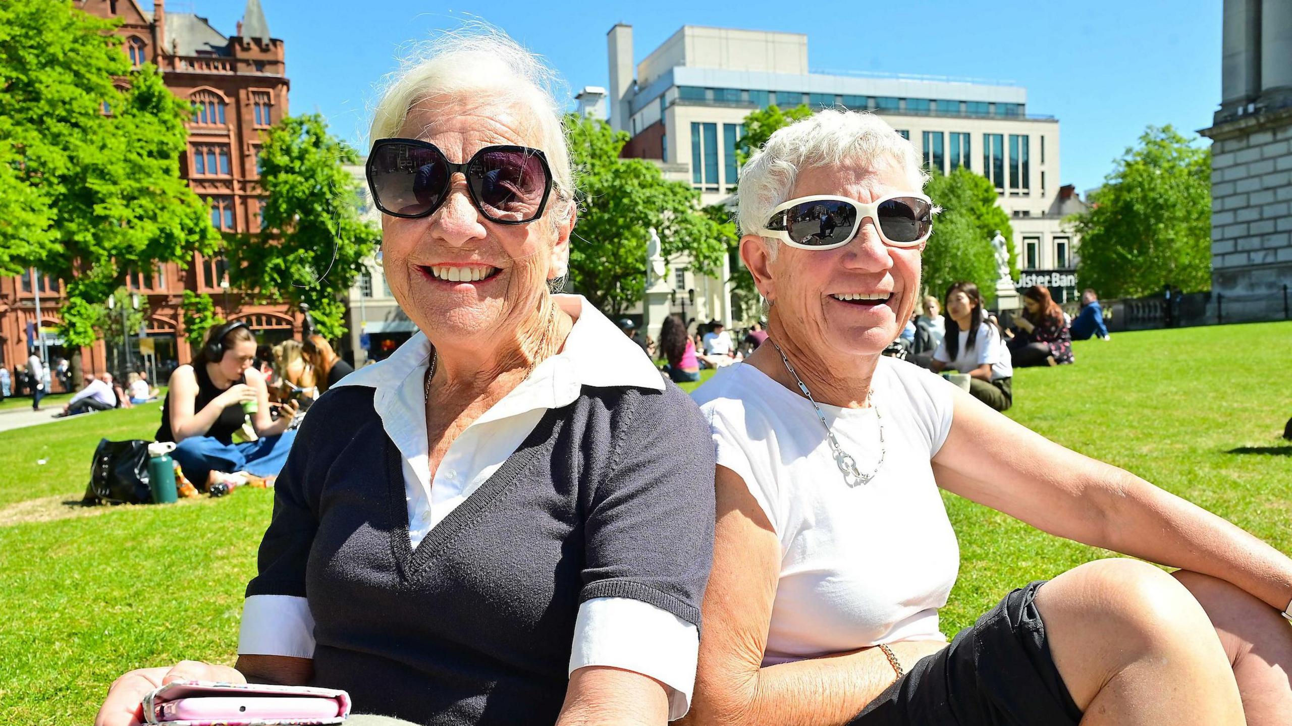 Sisters Jan Buchholz and Pam Sassella pictured sitting on the grass in the grounds of Belfast City Hall on 13 May 2025. They both have short, white hair and are wearing sunglasses and short-sleeved tops. They are sitting very close together and smiling at the camera on a bright, sunny day. Other people are sitting in groups behind them.