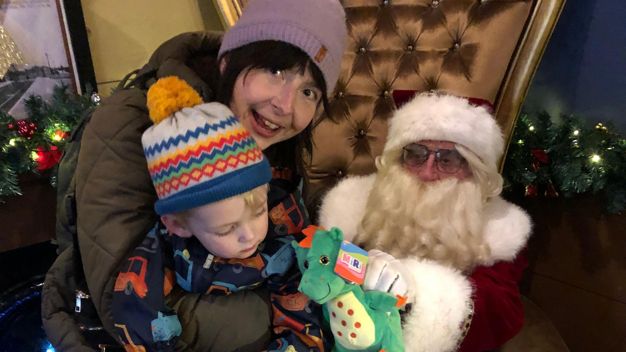 A woman wearing a coat and a woolly hat holding her young son, who is wearing a Christmas hat. He is holding a dragon toy that Santa has just given him. Father Christmas is on the right