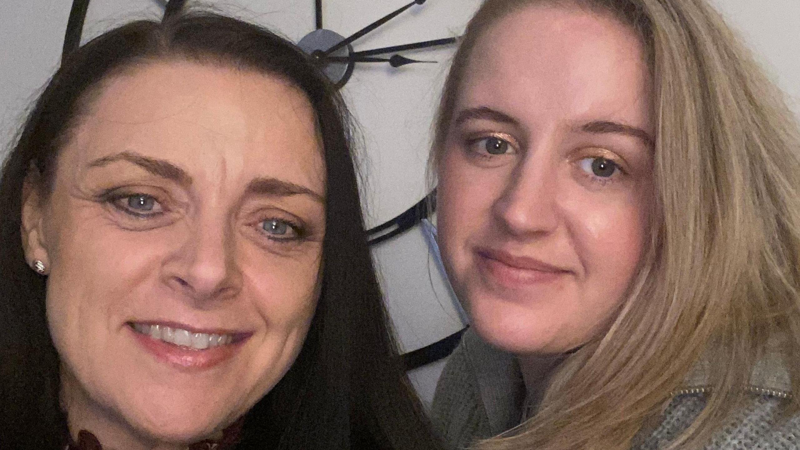 Abbi Smith and her mum, who has long brown hair and is smiling with a wall clock behind them both. Abbi is wearing a grey jumper and smiling.
