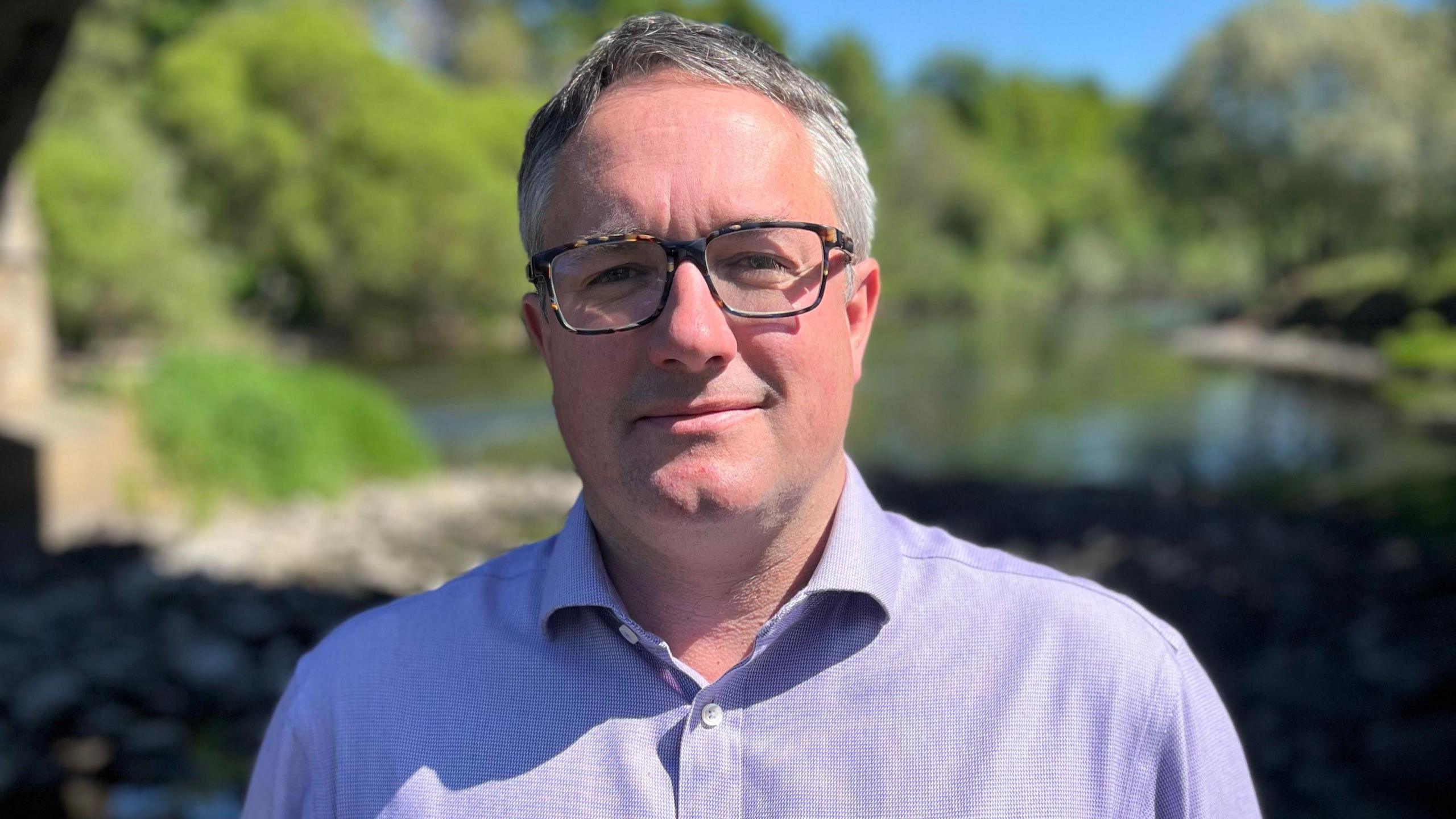 a man with grey hair and glasses wearing a purple shirt. He is standing in front of a small body of water which is surrounded by green trees.