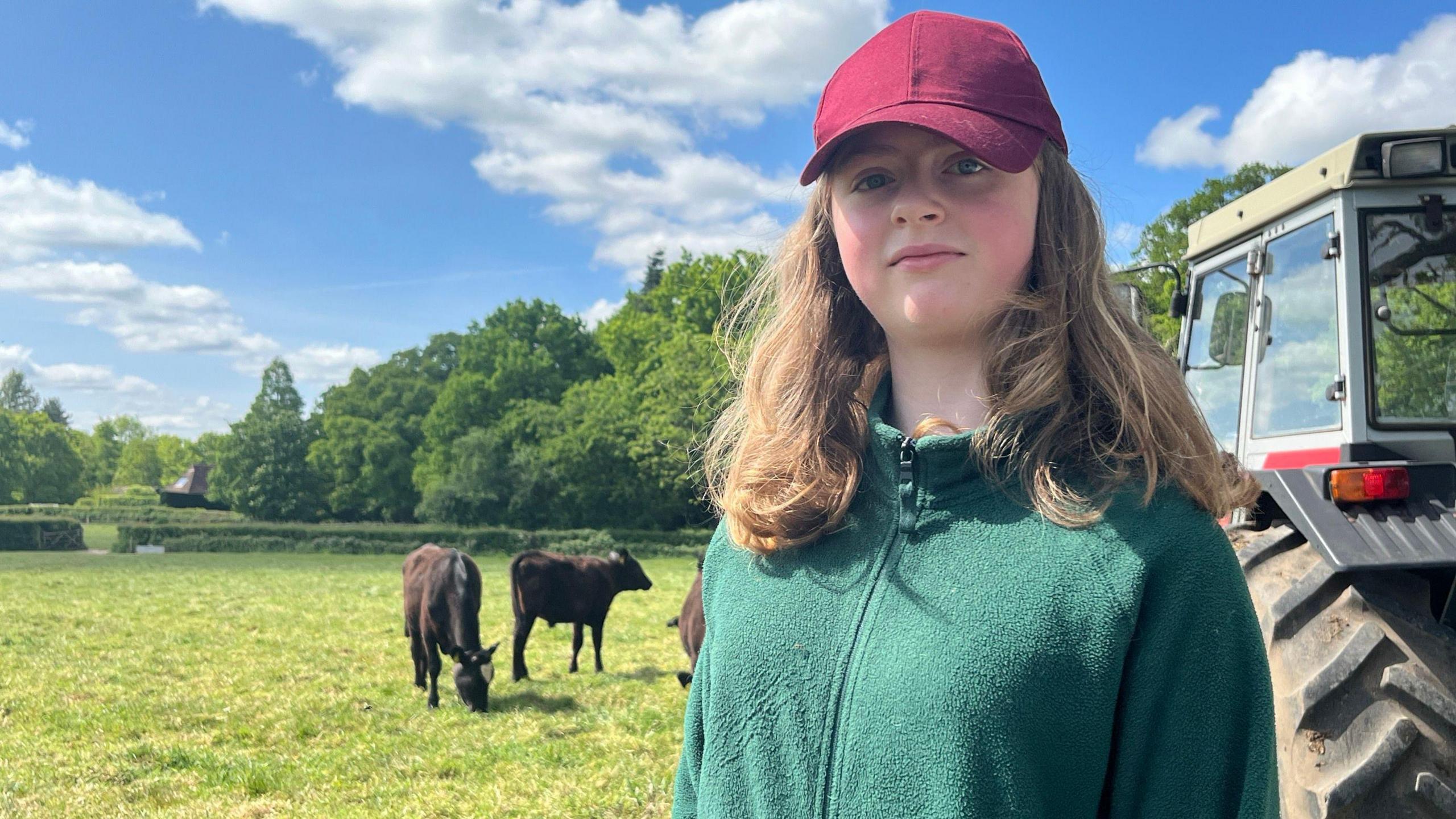 Dressed in a green top and red cap a teenager stands beside a tractor with large wheels. several cows are in the background and one is grazing the grass.