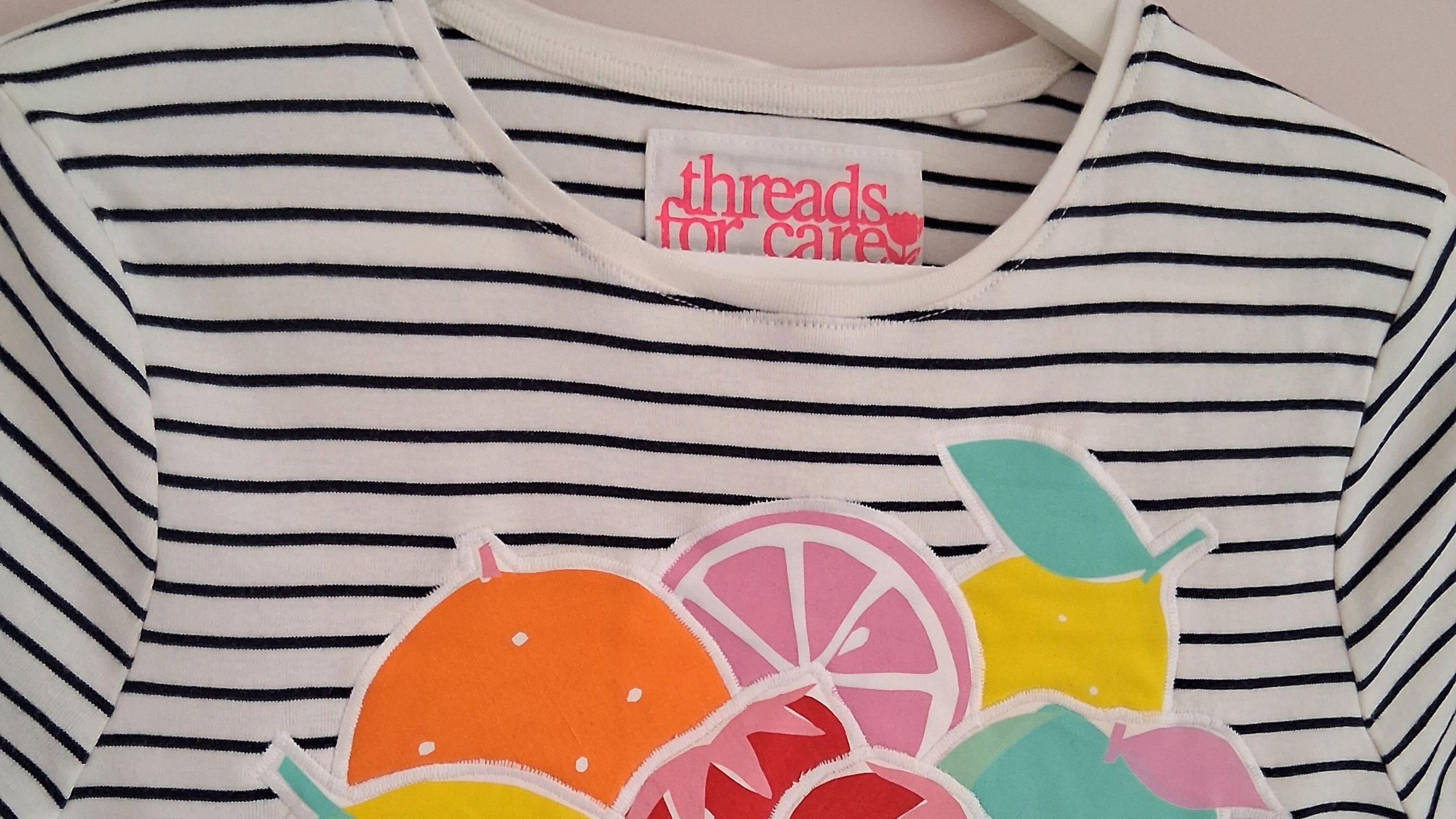 The top of a white a blue stripy t-shirt on a white coat hangar. It has had bright, cartoon fruit stitched on the front. A big threads for care label can be seen at the back