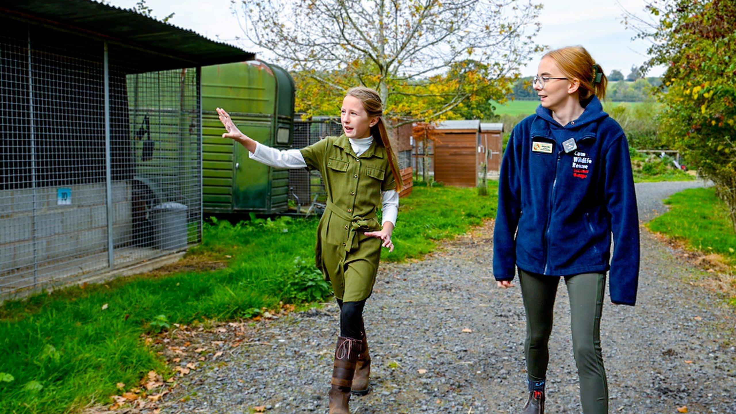 A young girl with light brown hair is wearing a green dress and white t-shirt and is walking on a stone path with her arm outstretched, talking. Walking next to her is a woman with auburn hair, wearing a dark blue jumper that has the cuan wildlife name on it. On their right are wildlife enclosures with wire netting in front of them