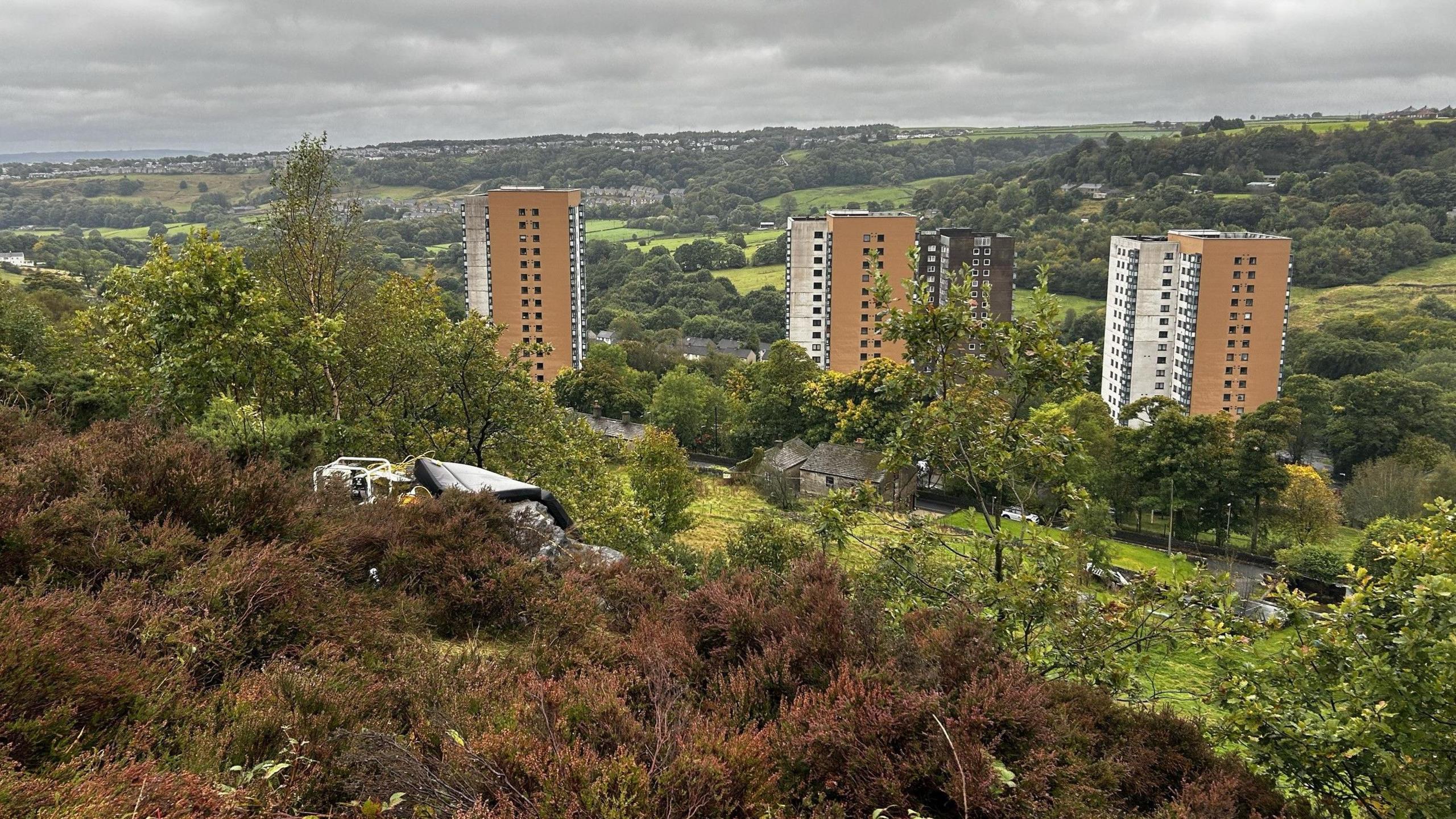 A cliff edge covered in heather, with some heavy machinery on the floor. Several tower blocks are situated down the hill among green fields and trees.