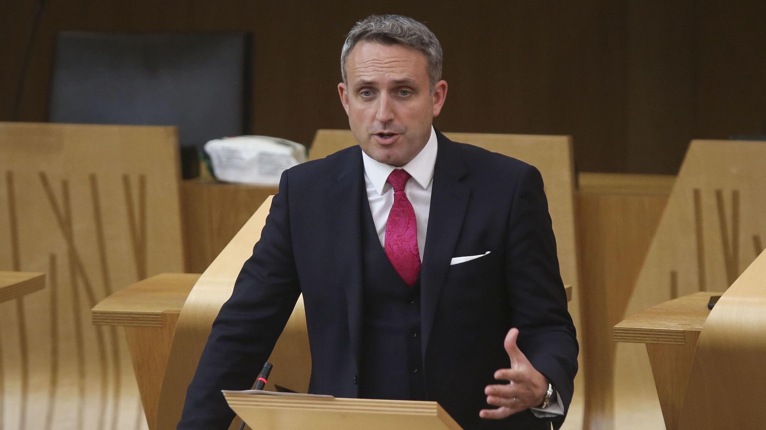 Alex Cole-Hamilton speaking in the Scottish parliament - he is wearing a suit with a purple/pink tie and has short, greying hair.