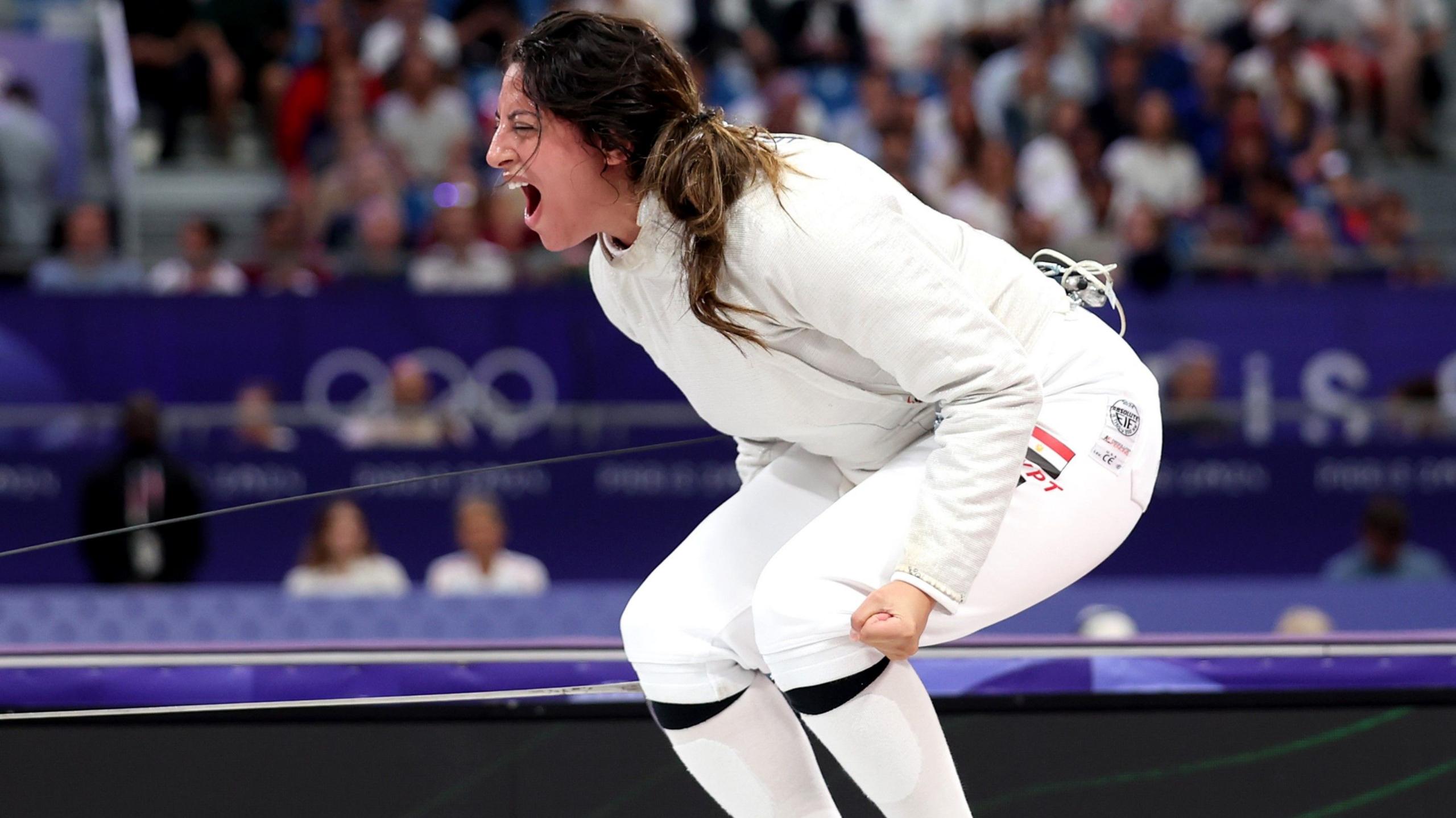 Nada Hafez, wearing a white fencing uniform, crouches down and clenches her left fist as she lets out a scream of celebration after beating Elizabeth Tartakovsky in the women's individual sabre event at the Paris 2024 Olympic Games