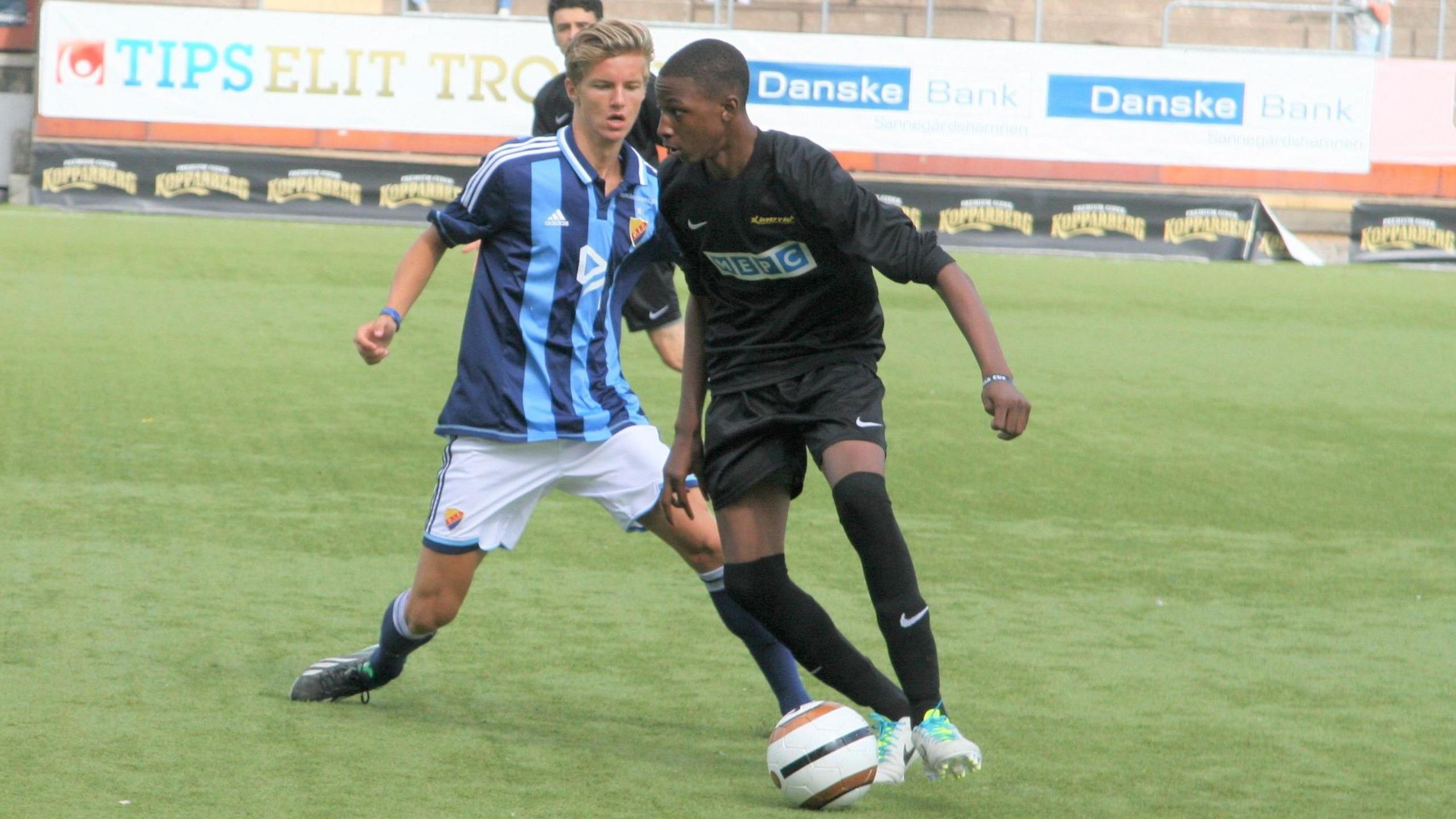A teenage Joe Aribo wears all-black kit as he controls a football with his left foot and, with head up, looks to turn away from an opponent in blue striped jersey and white shorts coming in to make a tackle from behind during a game for the Kinetic Foundation