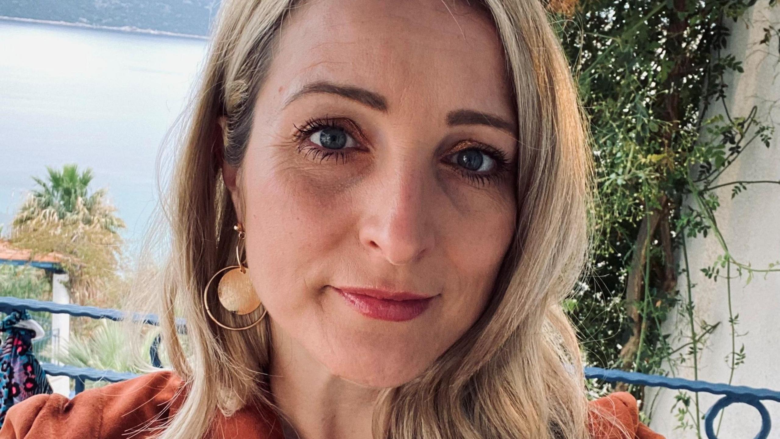 Portrait photograph of Jayne Clark, outside in front of the sea. She has blonde hair and is wearing gold earrings and a red top.