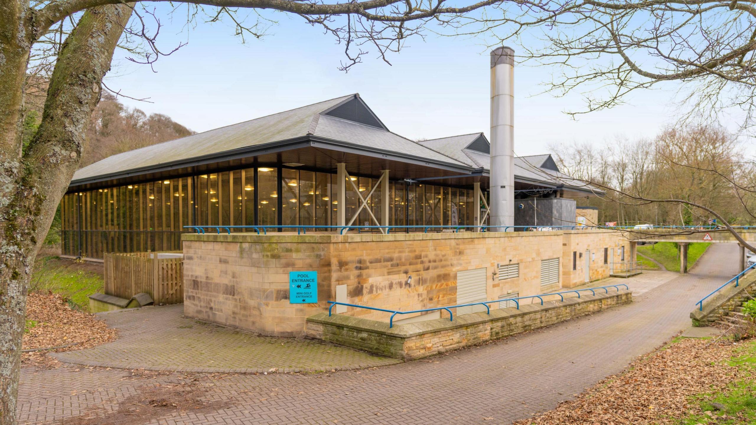 A modern building with a big glass front. A path snakes past it and a tree can be seen in the left corner.
