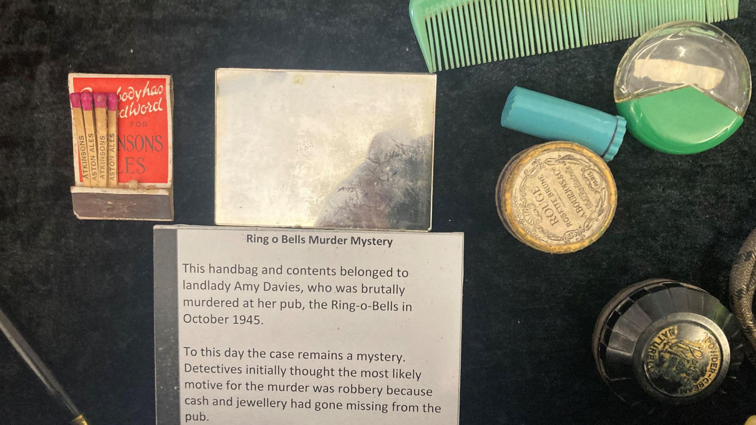 Make-up which belonged to Mrs Davis. There are several round pots and what appears to be a lipstick in a blue case. There are also a packet of matches in the photo and a sign with details of the "Ring O' Bells Murder Mystery". 