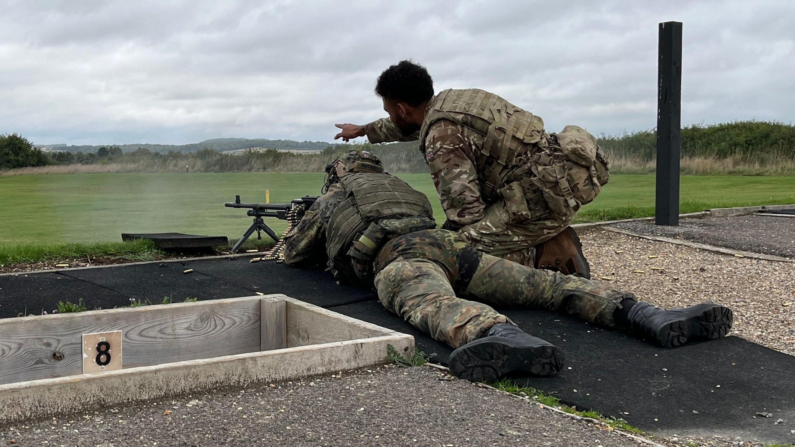 A soldier lying in a prone position shooting a mounted gun. Knelt over him is another solider pointing in a direction.