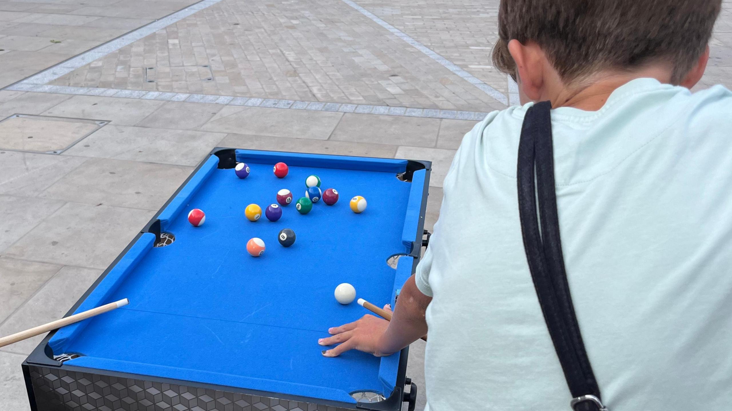A back view of a boy playing snooker on a mini pool table. He has short hair and is wearing a light green T-shirt. He is bent over the bright blue table, resting his left hand down, while the snooker cue rests between his fingers and thumbs as he gets ready to strike a white ball. A number of multi-coloured snooker balls are resting on the table. He is in Broad Street, March.