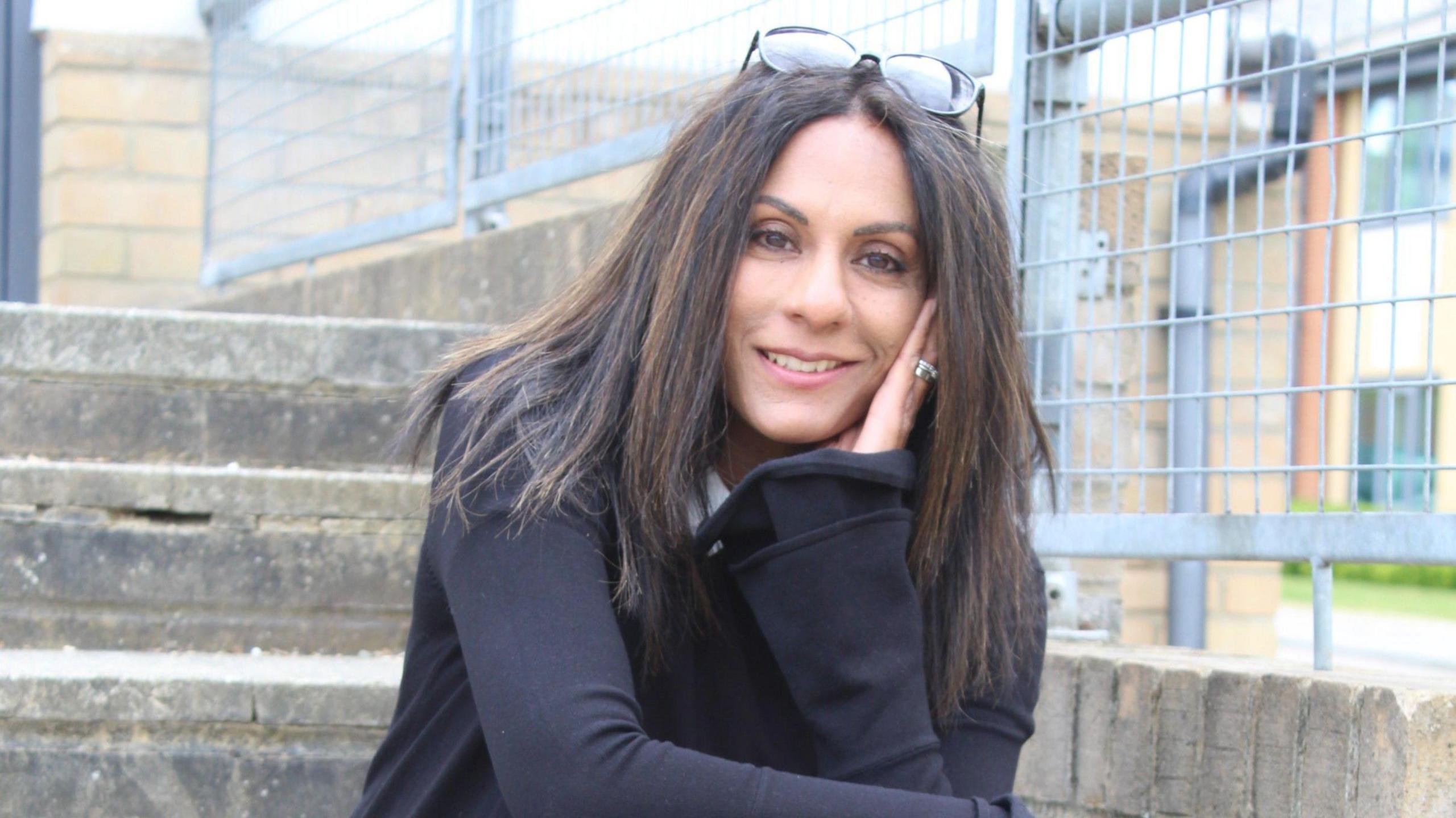 Kathryn Croft, a woman with long dark hair, wears a black jacket and poses on a set of concrete steps in front of a metal railing, her glasses pushed up on the top of her head, as she smiles into the camera.