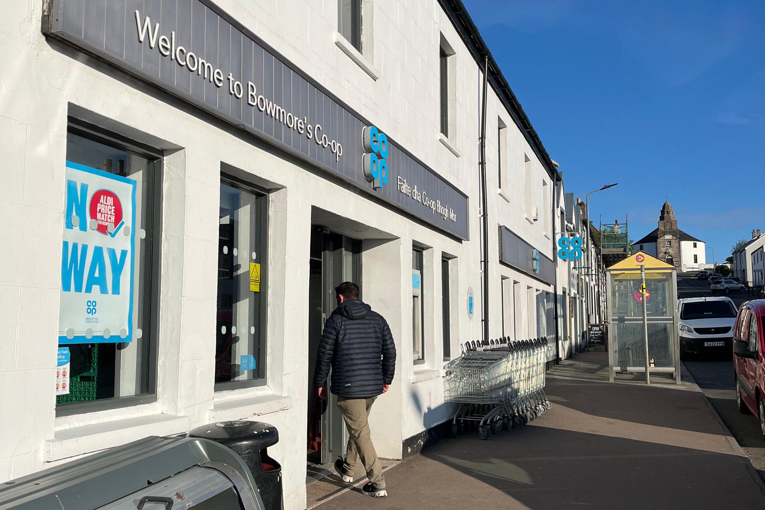 The front entrance of a Co-op grocery shop in Bowmore on Islay. A man wearing a blue puffer jacket and light chinos, with his back to the camera, walks in through the doorway.