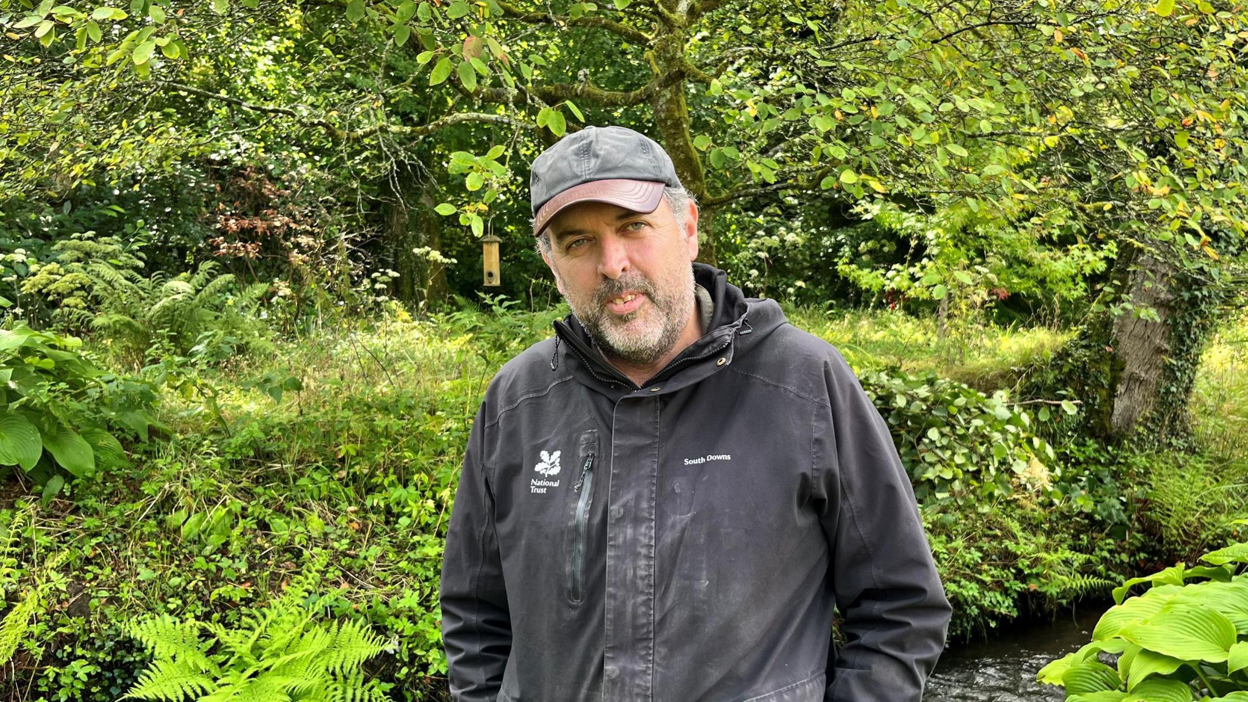 A man wearing a black coat and grey cap. The coat has a leaf logo on it with the words "National Trust" on the left hand side. He is stood in a green, leafy area near a body of water.