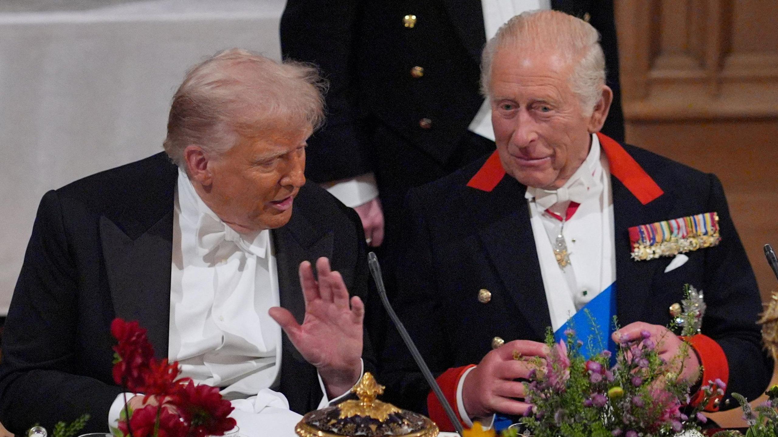 King Charles III and US President Donald Trump sit down next to each other in front of a table with gold ornaments and flowers.