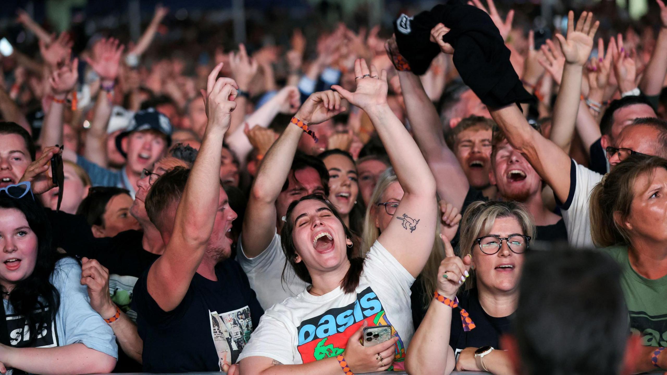 Fans can be seen singing with their arms in the air at the front of the crowds of the Oasis gig in Cardiff. One girl, centre, wears an Oasis merch t-shirt and has her mouth wide open singing. 