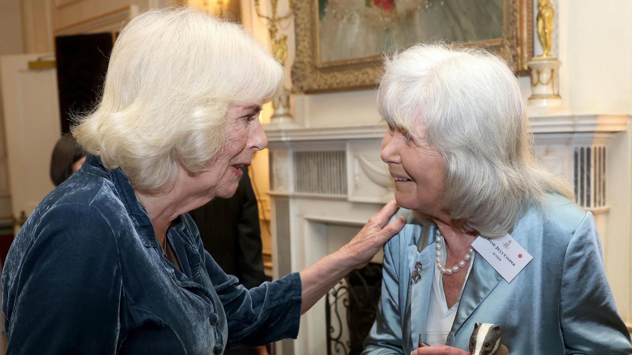 Queen Camilla and Dame Jilly Cooper speaking at a reception at Clarence House, London, in March 2025, and the Queen has one hand on Dame Jilly's shoulder