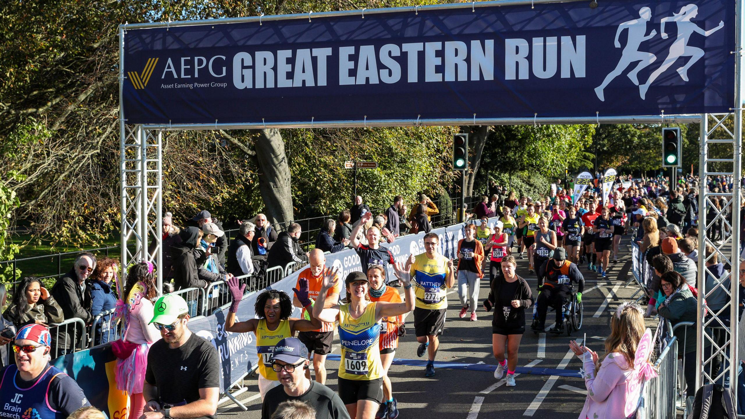 People in colourful T-shirts running and waving to the camera with a blue banner saying Great Eastern Run 