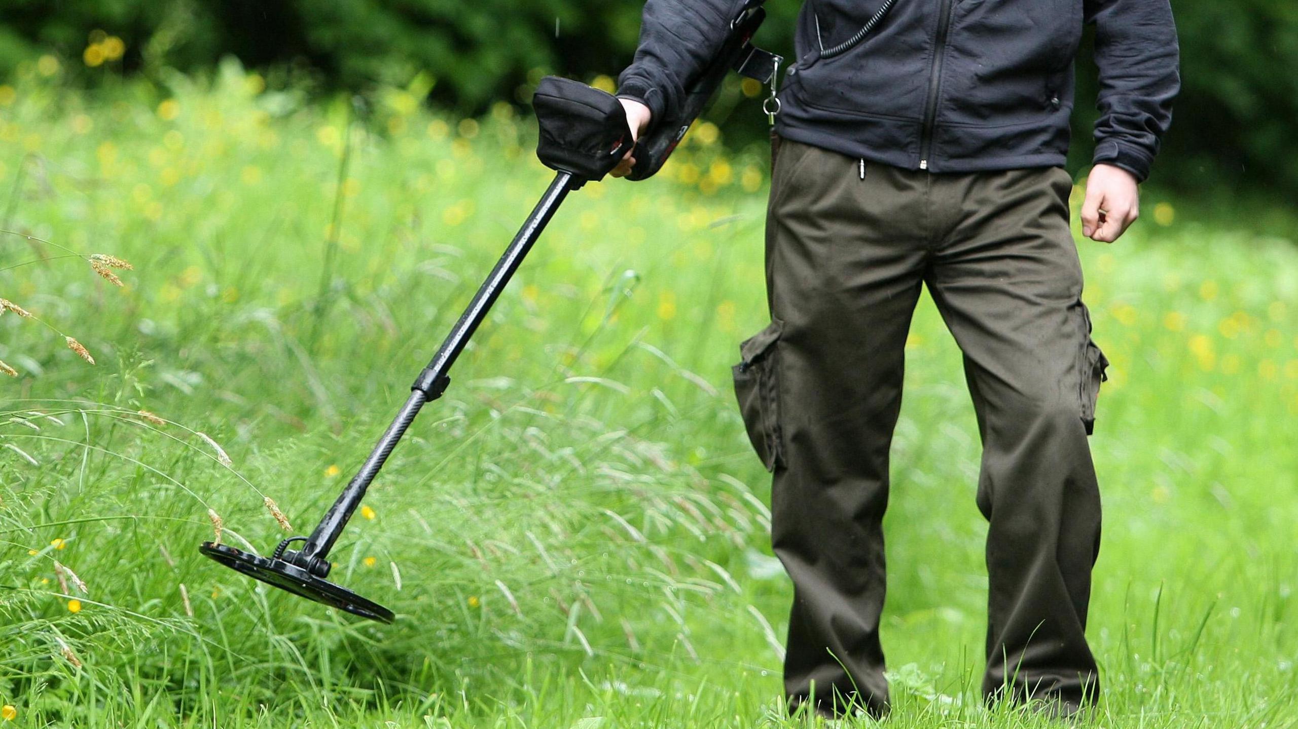 Generic photo of a metal detectorist scanning a grassy area. The detectorist is wearing dark green cargo pants.