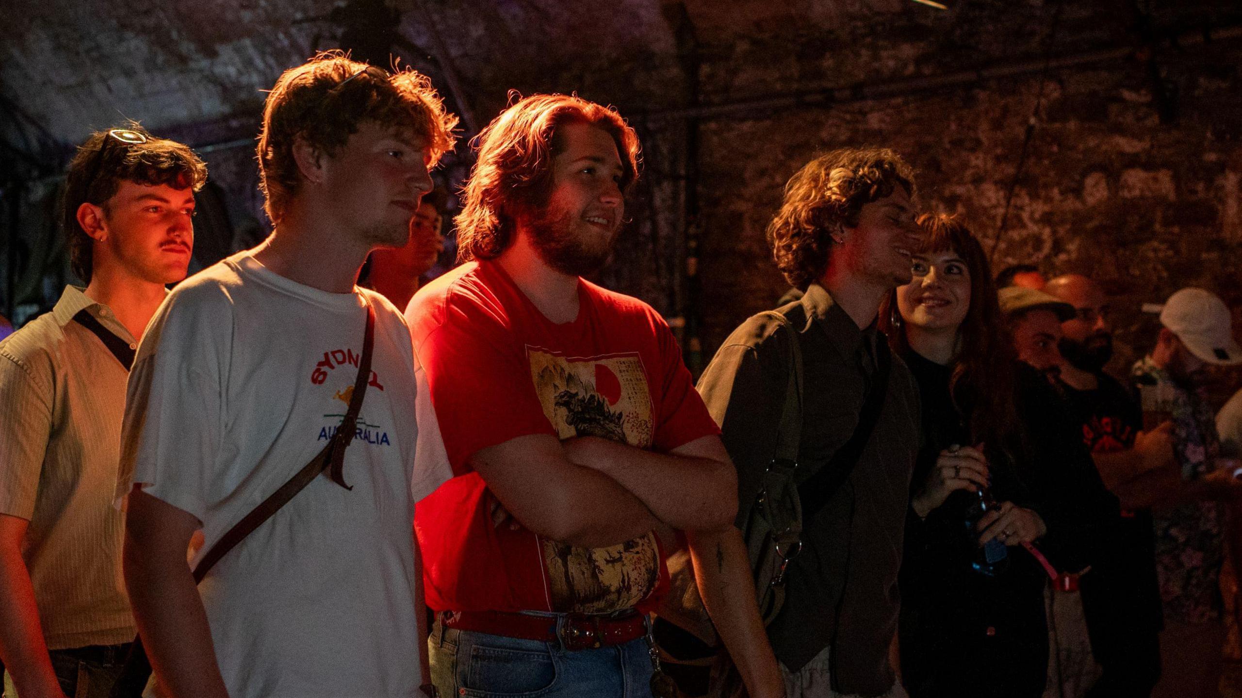 A group of young people watch an exhibition of musical instruments and technology at dBs Institute of Sound & Digital Technologies in Bristol. They are illuminated by red light and are in a room with dark stone walls