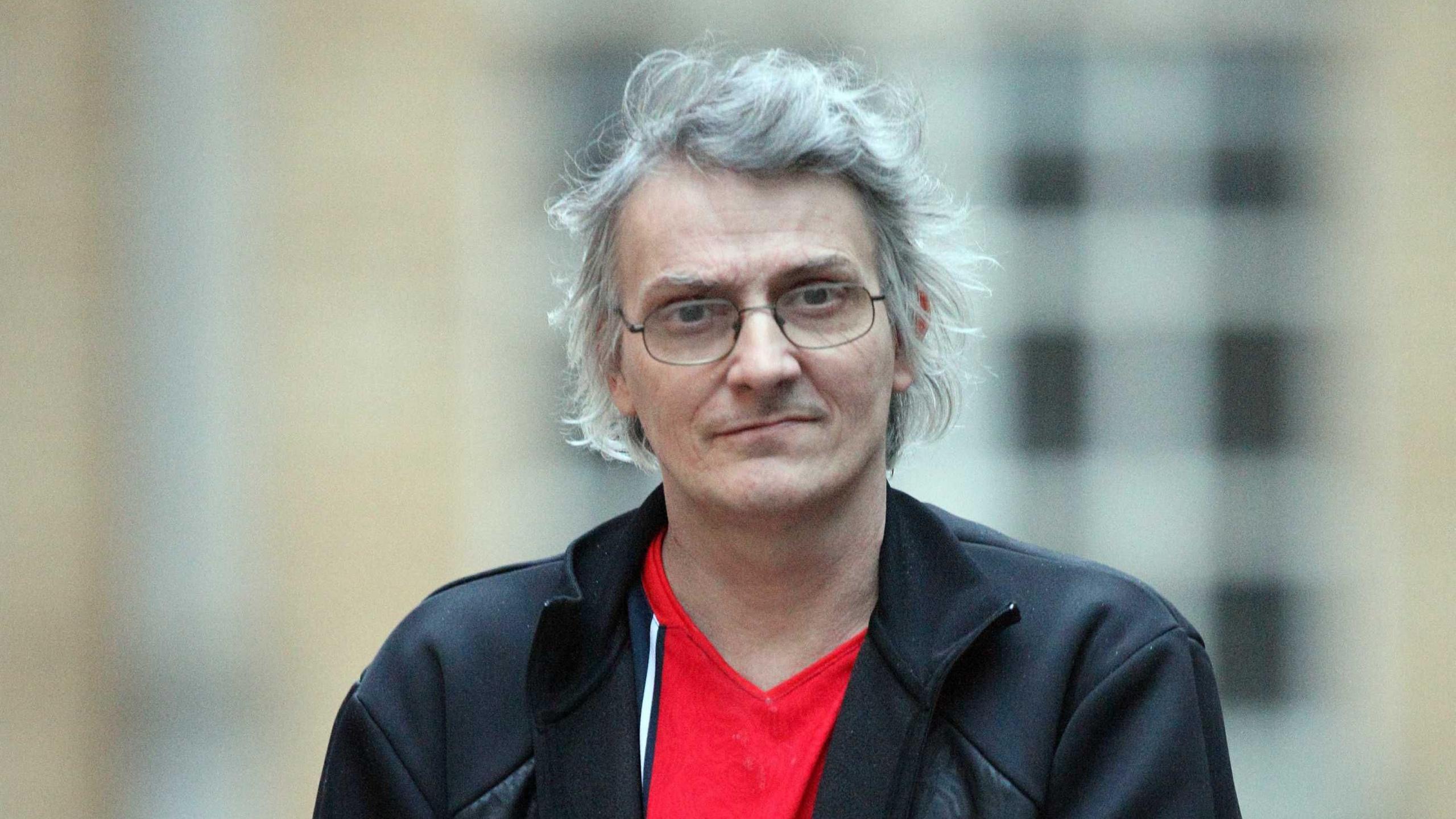 A man is looking at the camera. It is a head and shoulder shot. He is wearing black thin-rimmed glasses and has grey/silver hair