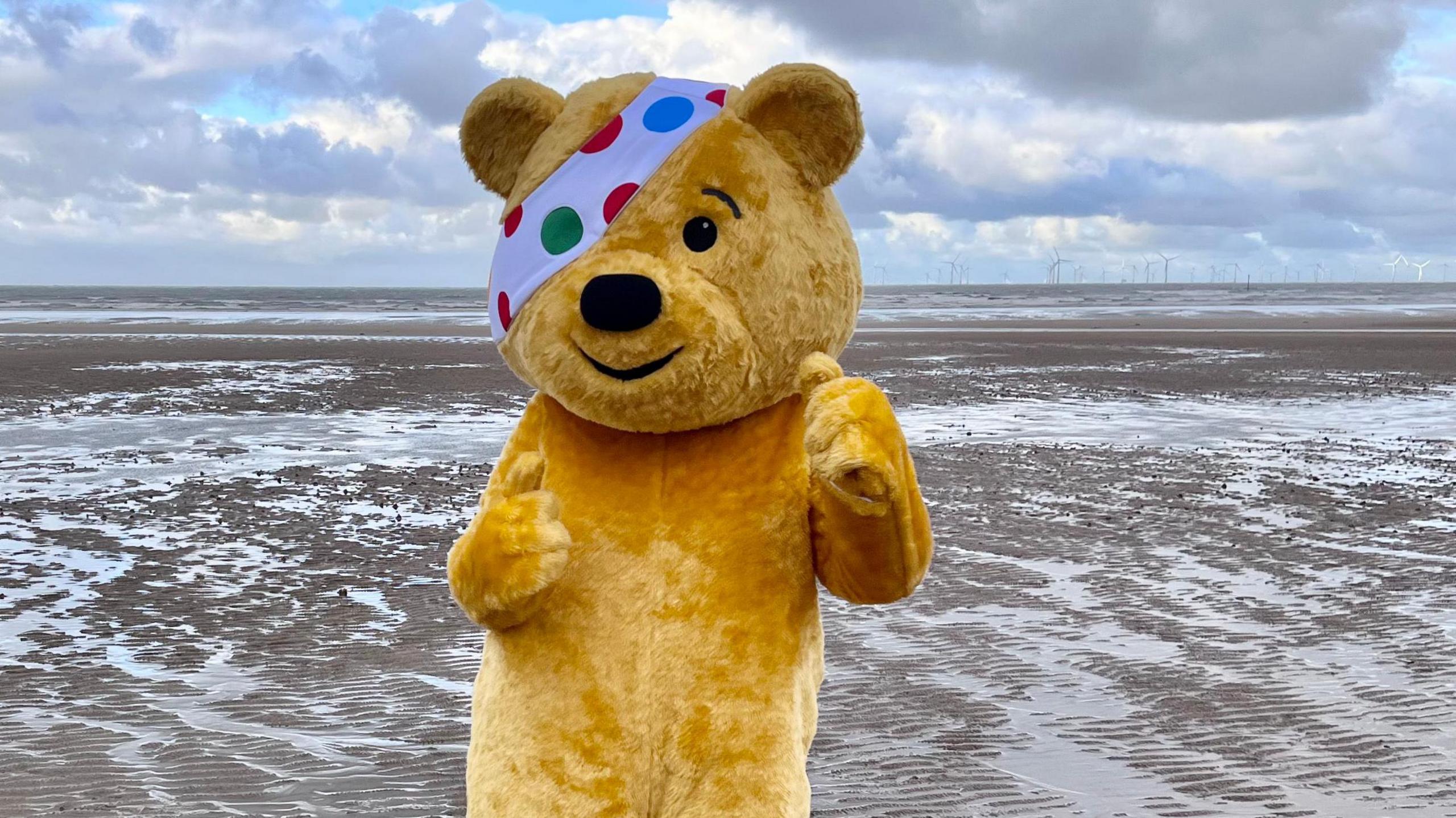 Pudsey the Bear stands on a beach. He is a large golden anthropomorphic bear and is wearing an eye patch which is white with green, red and blue spots on it.