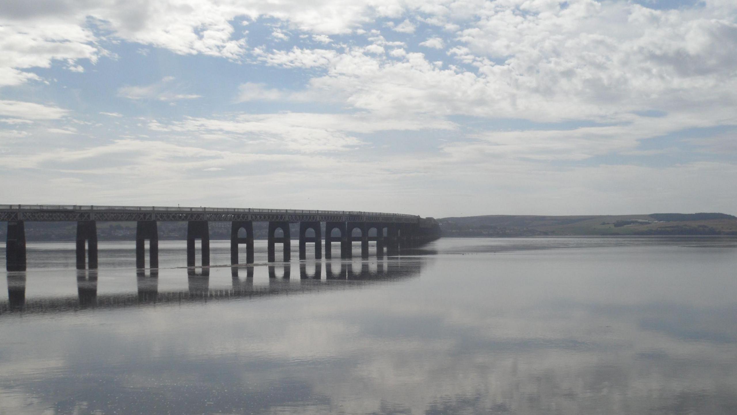 A long bridge stretches across a wide estuary under a partly cloudy sky. The still water mirrors both the bridge and the clouds.