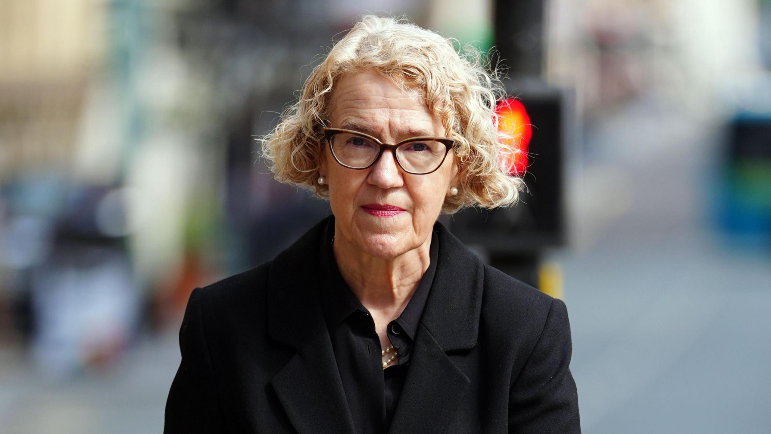 Chair of the inquiry Lady Justice Thirlwall looks at the camera. She is wearing a black smart jacket. She has short curly hair and is wearing glasses. 