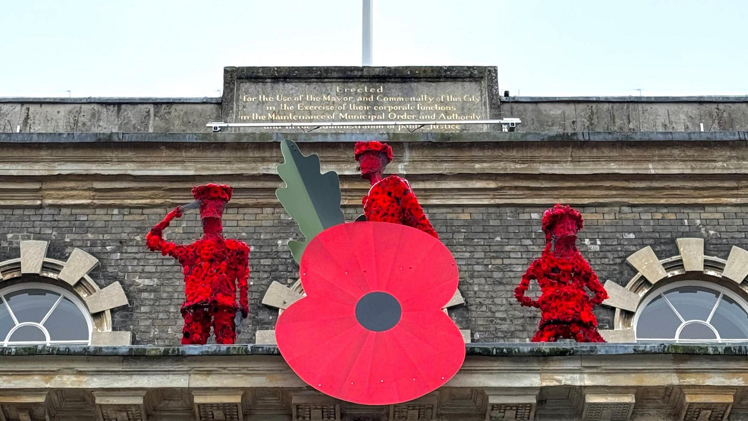 The top of a grand building under a grey sky - a huge version of a Royal British Legion poppy is attached to the front and behind it are three soldier-type figures which are covered in knitted poppies.